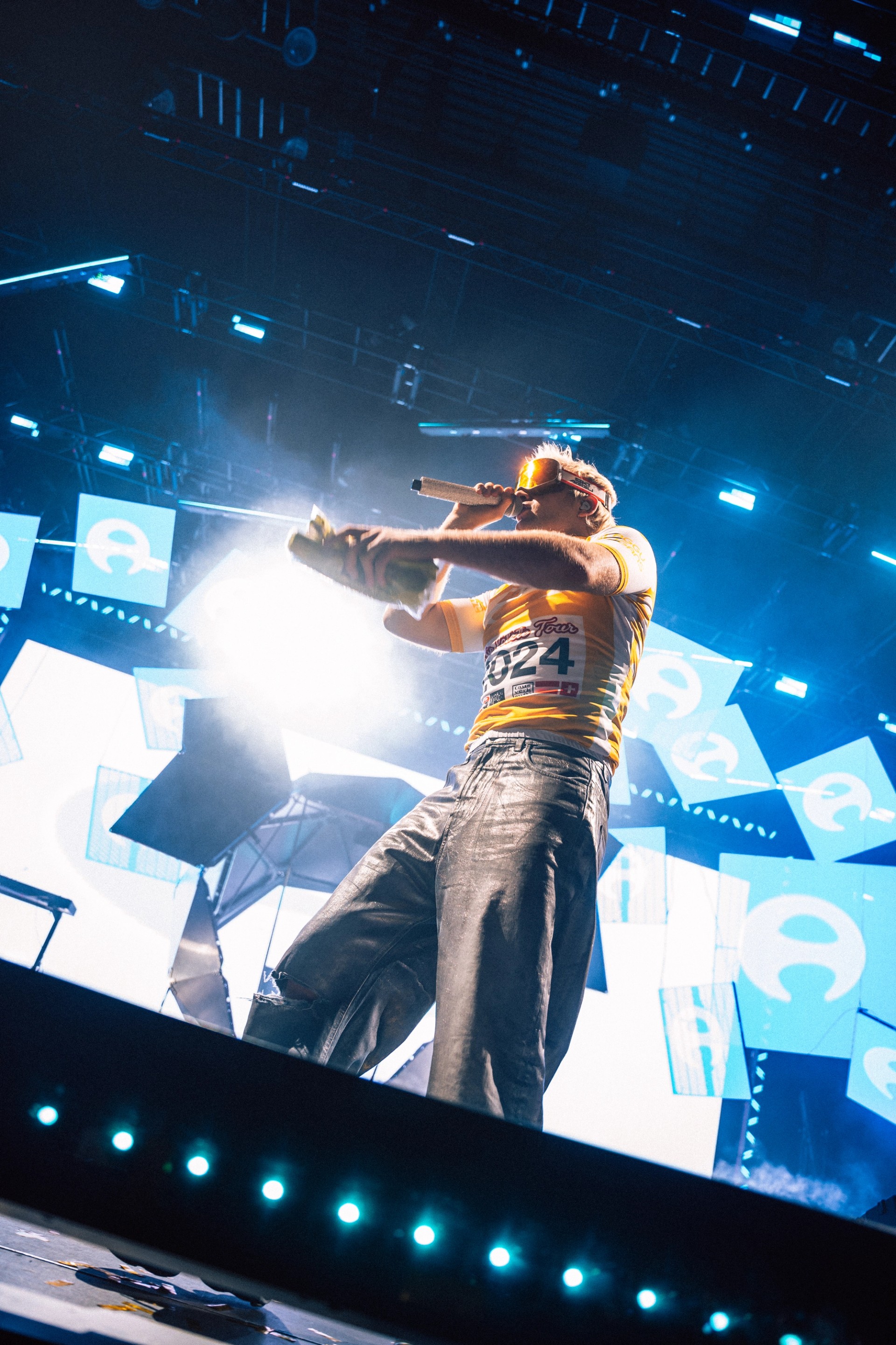 Low-angle shot of Ski Aggu pointing into the crowd in a yellow jersey, in front of tilted LED panels showing a white logo on blue.