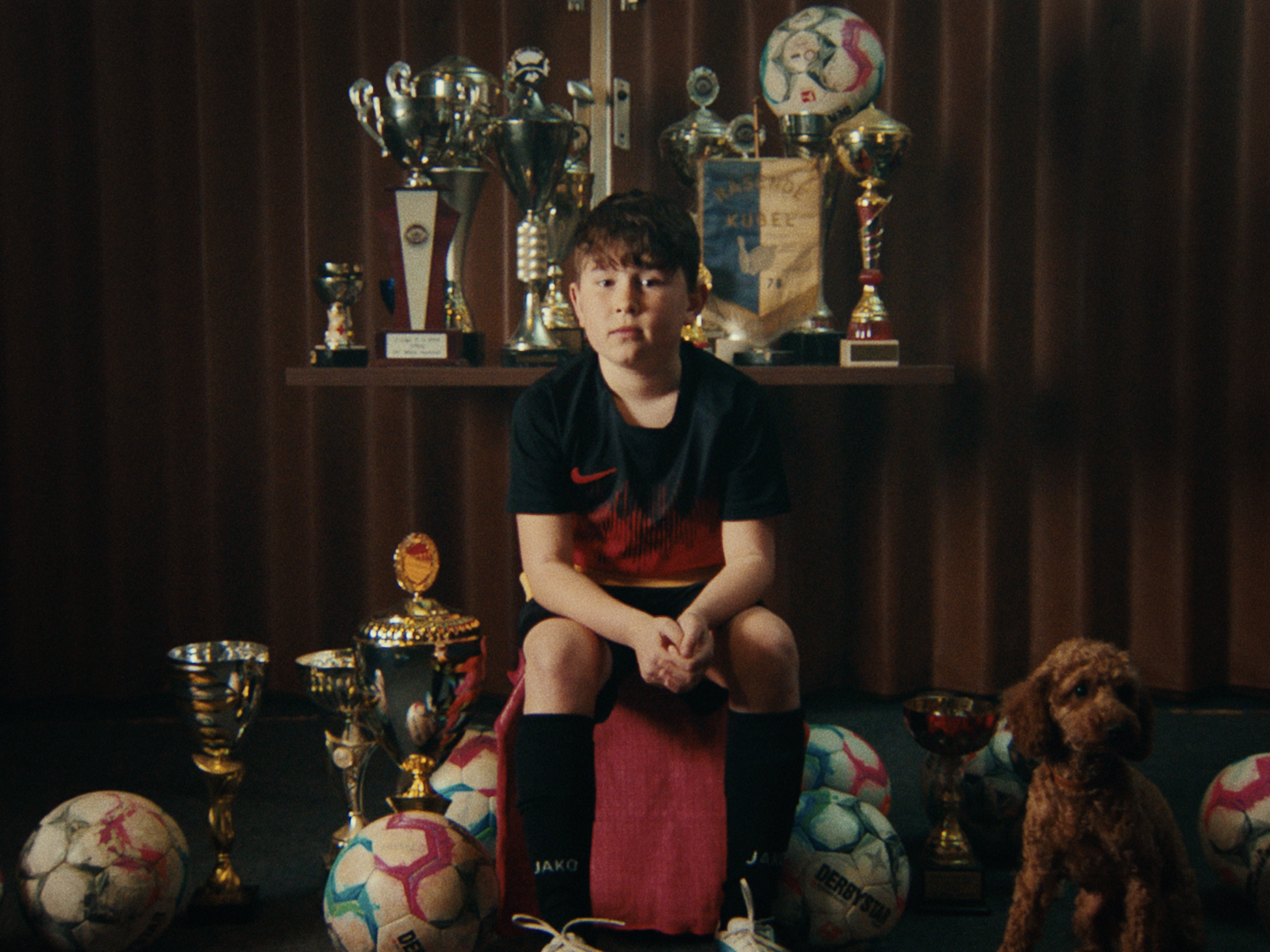 Boy in a football shirt sits among trophies and footballs in front of a brown curtain, a small brown dog standing beside him.
