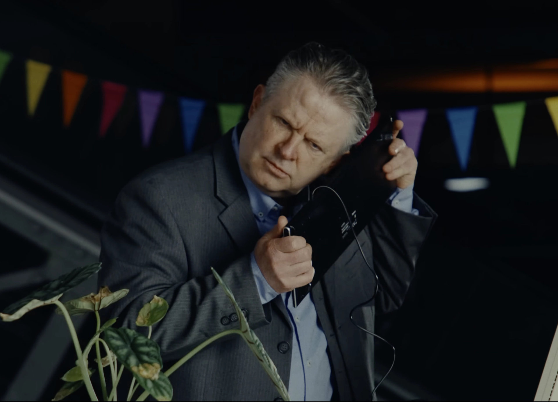 The car salesman in the grey suit frowns as he presses a black keyboard with a cable to his ear like a phone, bunting behind him.