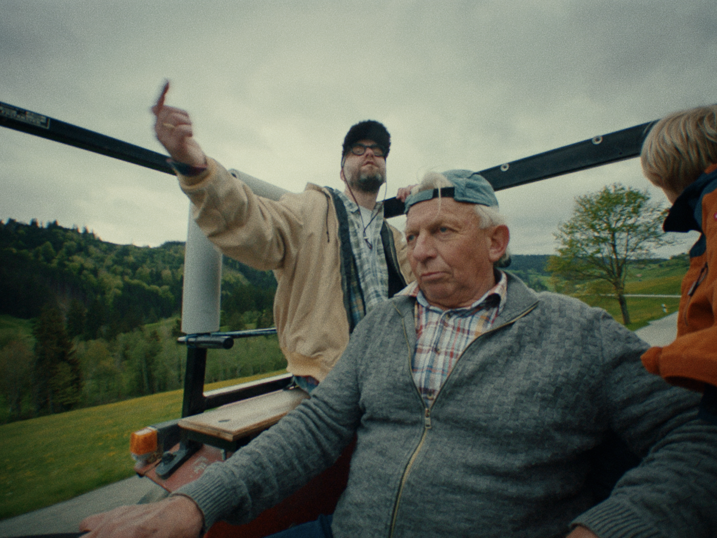 On the back of a tractor, Mark Forster in a cap and beige jacket points ahead behind the older driver in a grey cardigan, wooded hills around them.