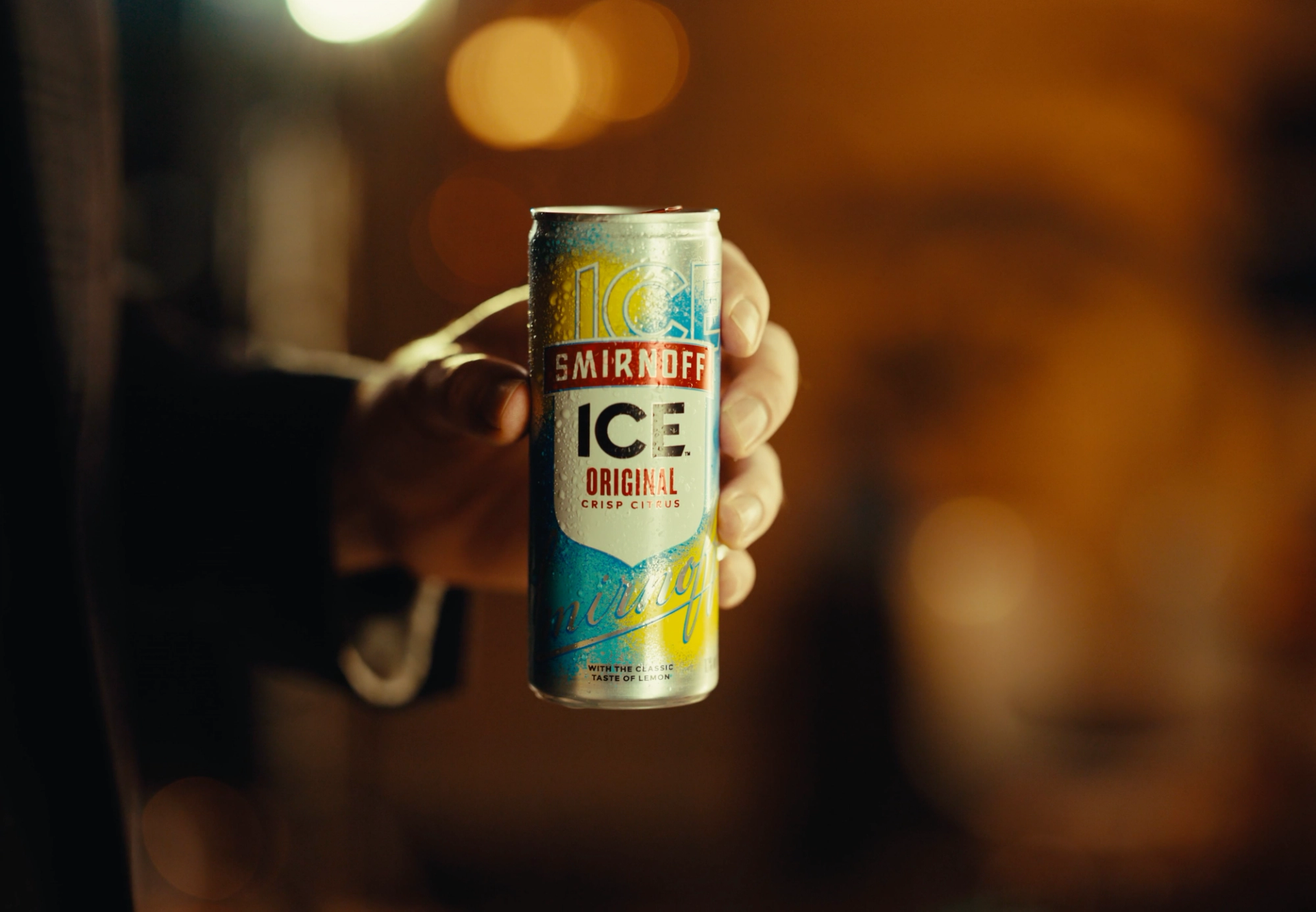 Hand holding a wet can of "Smirnoff Ice Original" towards the camera, with warm out-of-focus lights behind.