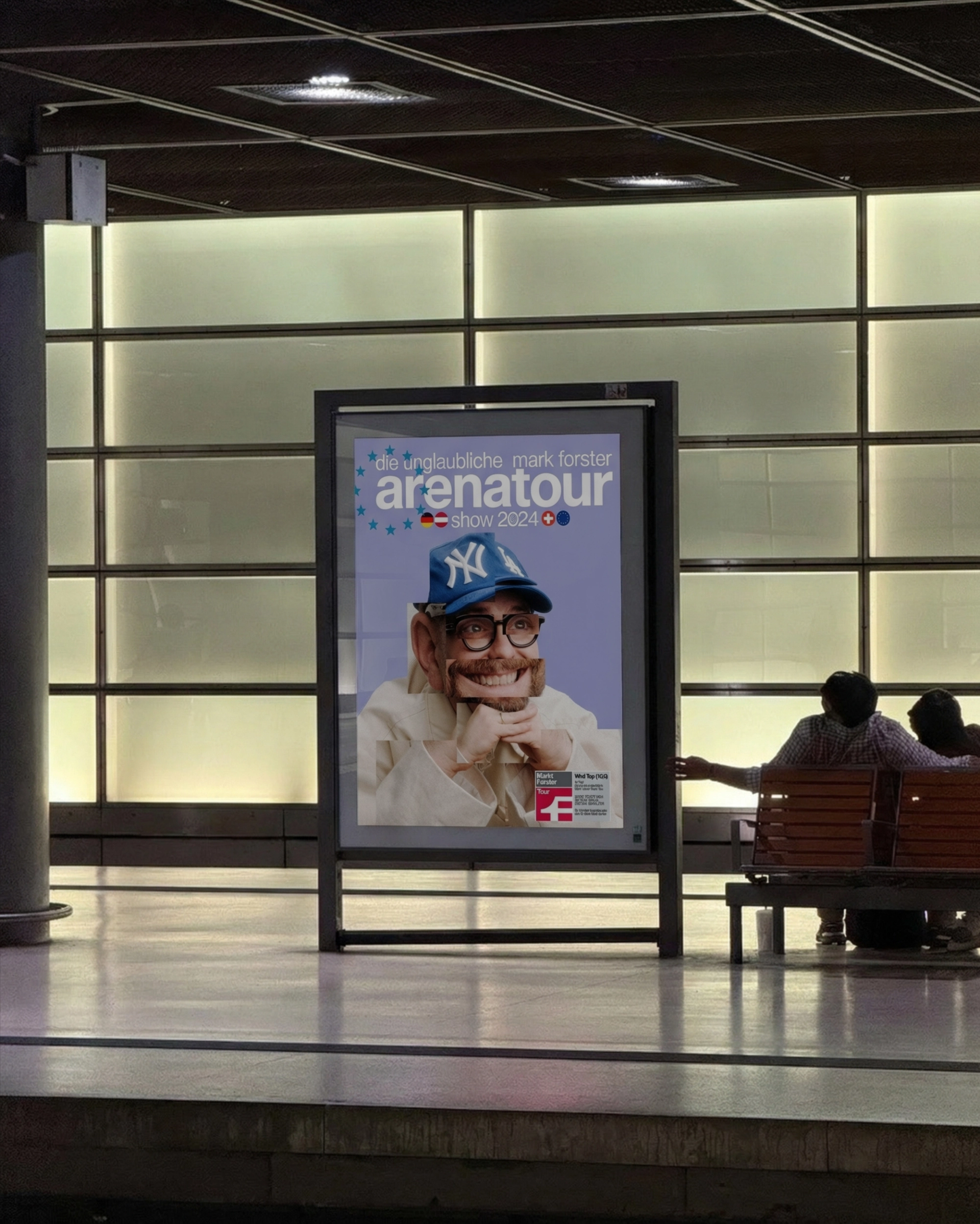 The arena tour poster in a backlit display on a station platform at night, two people sitting on a bench next to it.