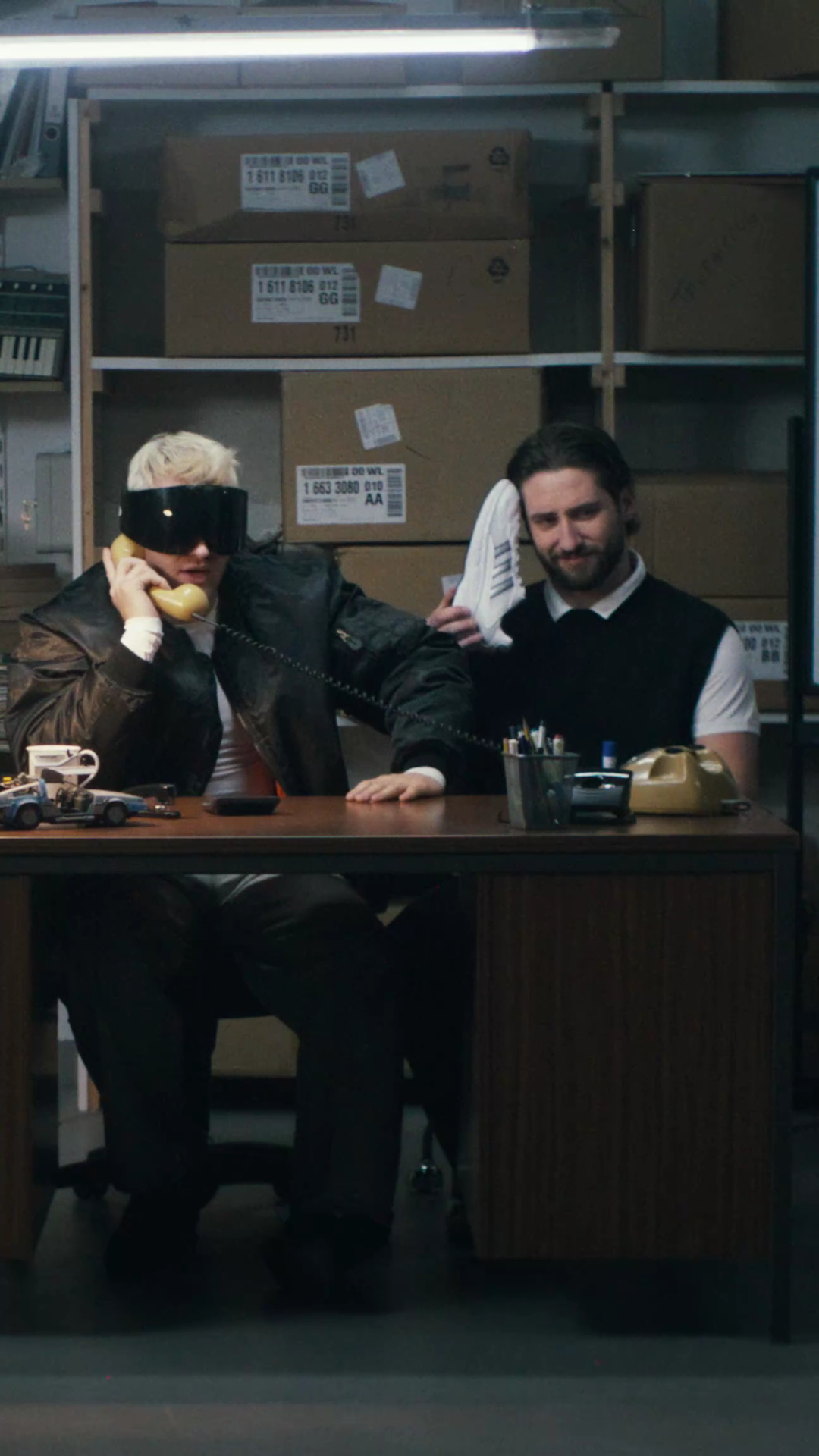 Stockroom scene in upright format: Ski Aggu on a yellow rotary phone at a desk while a bearded man next to him holds a white sneaker to his ear like a receiver.