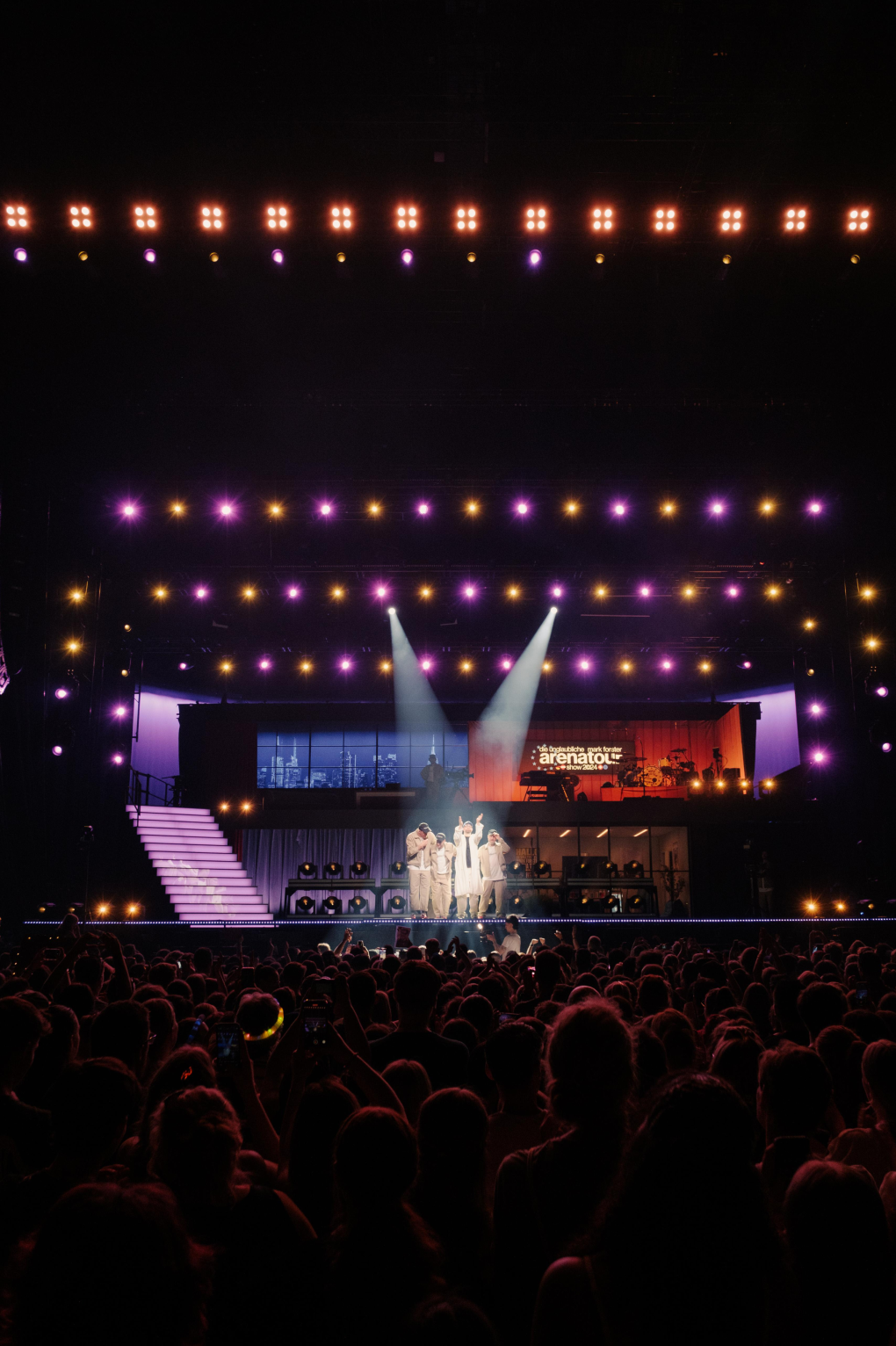 Four performers under two crossing spotlights at the front of the stage, seen from the middle of the crowd.