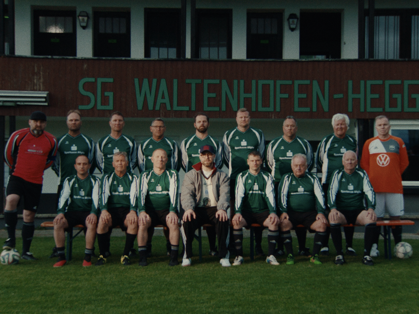 Team photo in front of a clubhouse lettered "SG WALTENHOFEN": footballers in green kits in two rows, Mark Forster in a cap and track jacket seated in the middle.