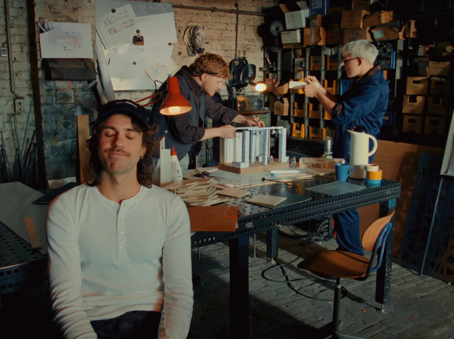 Workbench in the workshop: two band members assemble a small white model under an orange lamp while the third sits in front with his eyes closed.