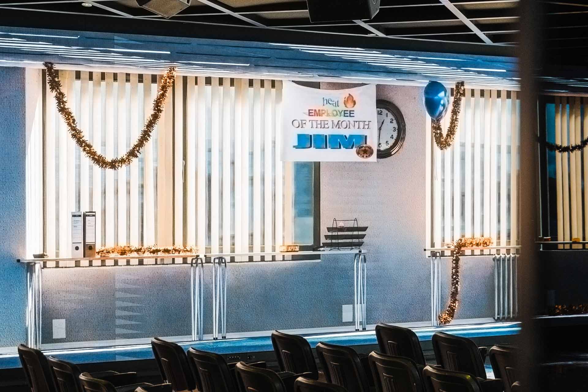 Screen wall behind the seating with an "EMPLOYEE OF THE MONTH" banner, a wall clock, gold tinsel and a blue balloon in front of vertical blinds.
