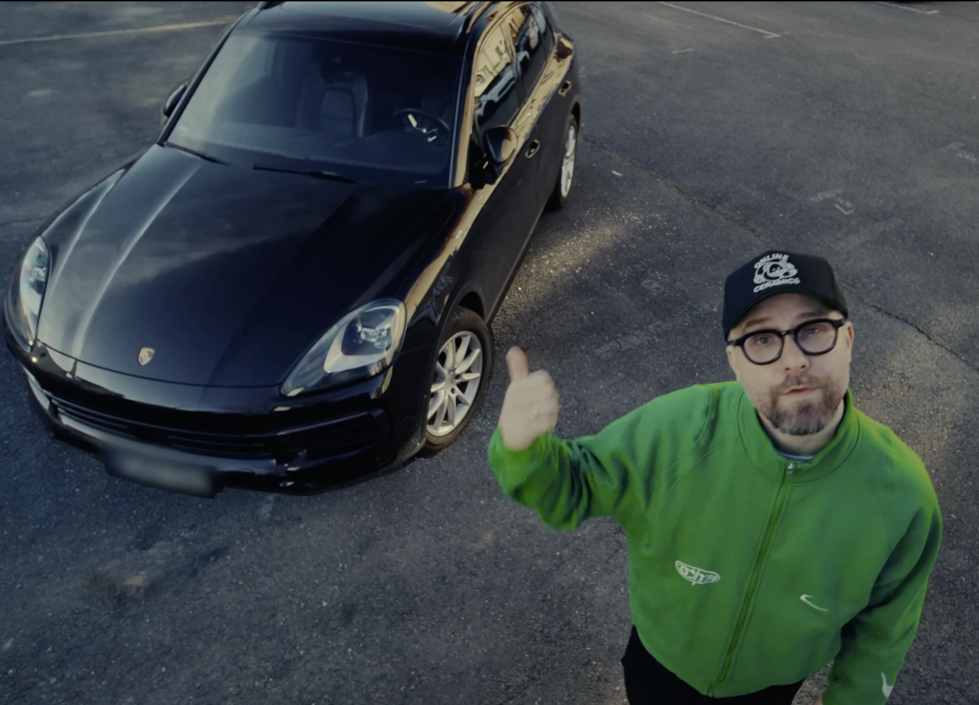 The same thumbs-up from Mark Forster in the green track jacket, this time next to a black Porsche SUV, in a tighter crop from above.