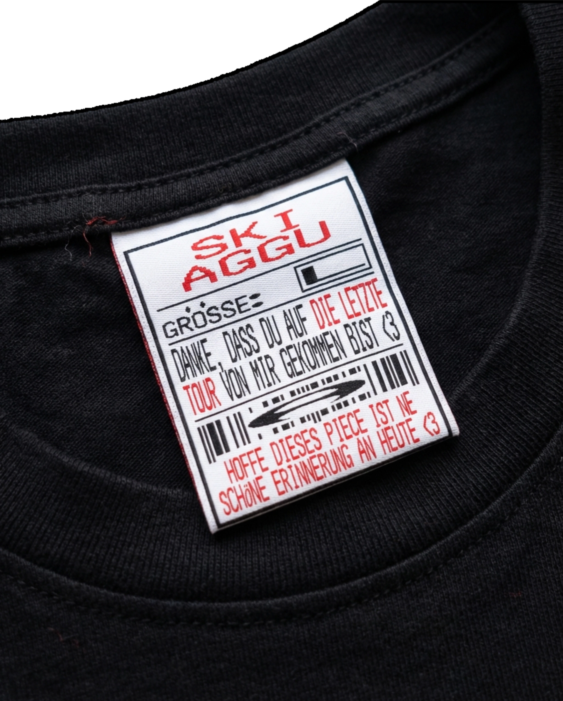 Woven neck label in a black T-shirt: "SKI AGGU" in red above a size field, barcodes and a thank-you note for coming to the last tour.