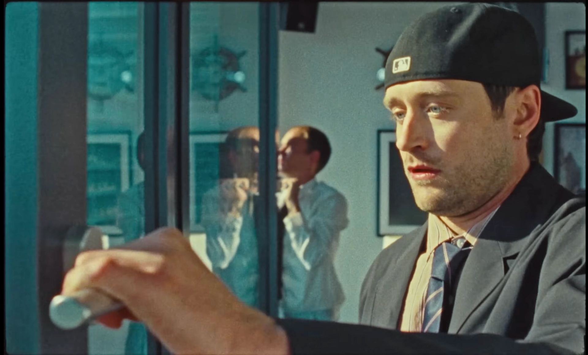 Close-up of Ritter Lean in the backwards cap reaching for a door handle at the window, with Flaschko in a white shirt out of focus behind the glass.