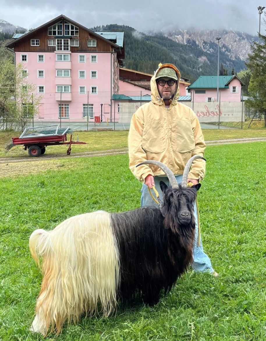Mark Forster in a pale yellow hooded jacket holds a long-haired black-and-white goat on a lead in a meadow, a pink alpine hotel and mountains behind.