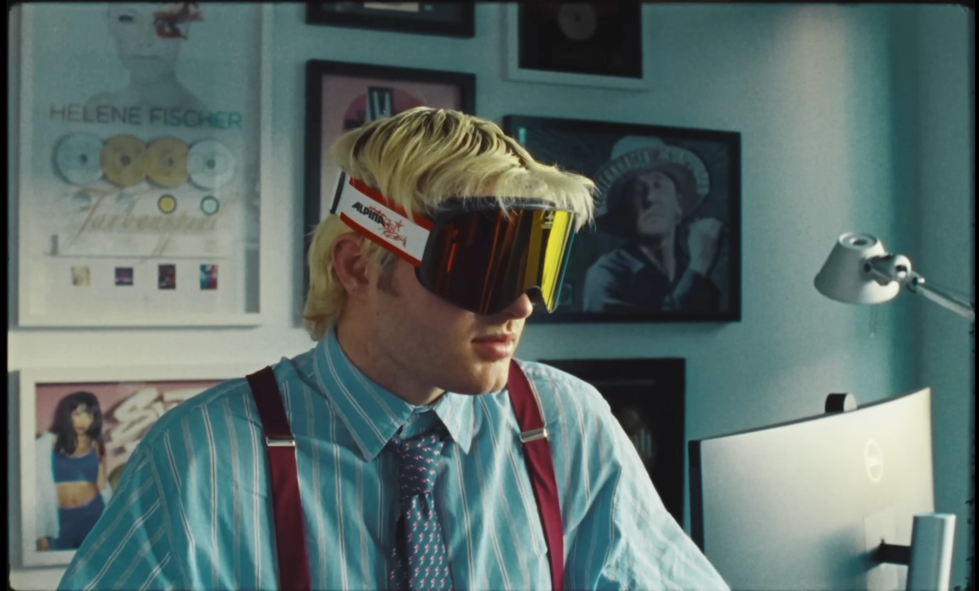 Ski Aggu at a desk in a striped shirt, red braces and ski goggles, in front of a wall of framed gold records.