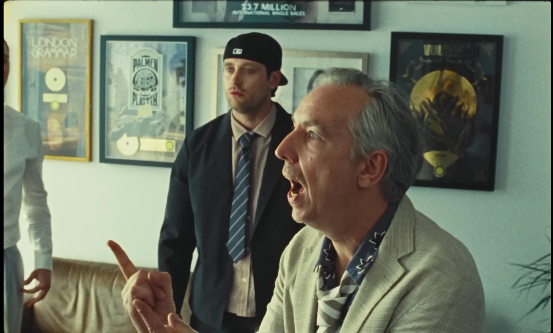 Olli Schulz, grey-haired in a light blazer, argues with a raised finger while Ritter Lean stands behind him in front of framed records.