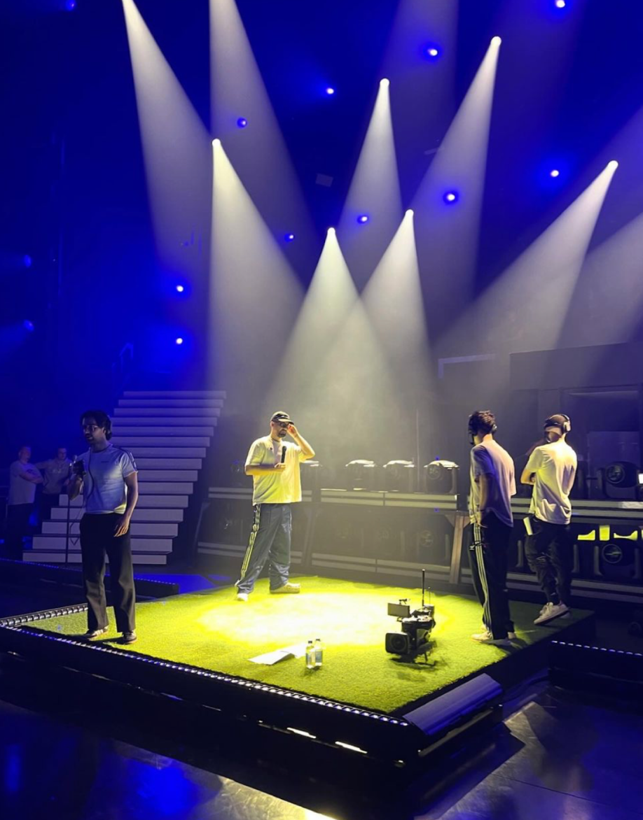 Stage rehearsal: four people on a square platform covered in artificial turf, one holding a microphone, under white spotlights and blue light with white stairs behind.