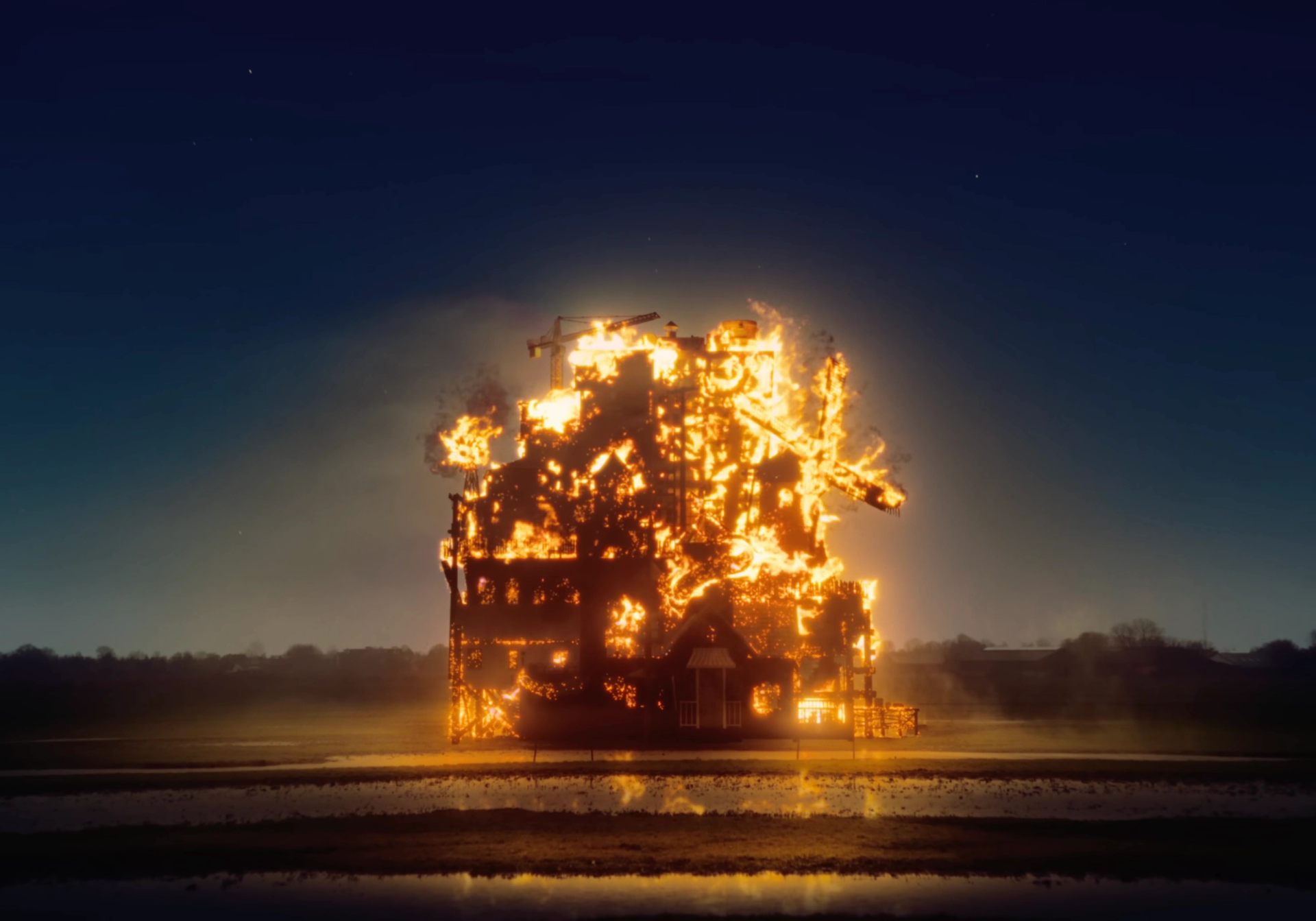 The whole house burning at night under a starry sky, the flames reflected in the flooded field in front of it.
