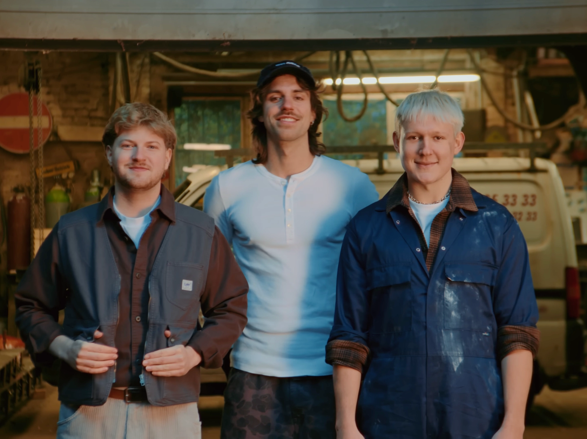 The three members of 01099 in work clothes, one in paint-stained blue overalls, smiling in front of the workshop and a white van.