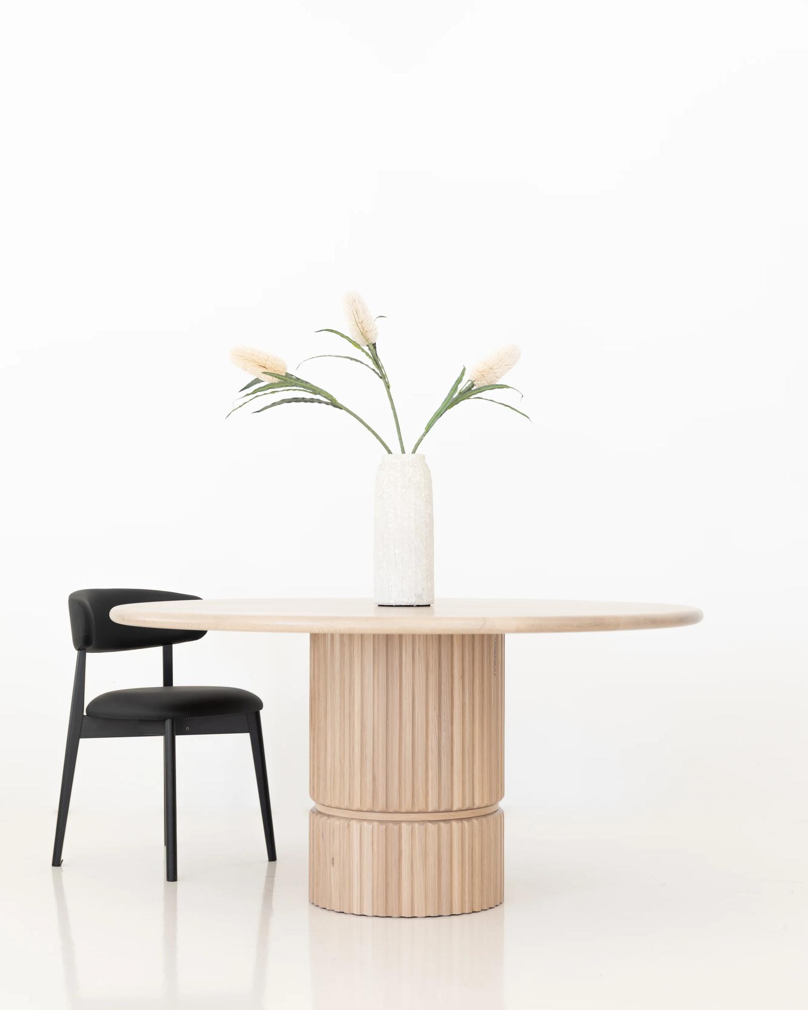 Esto Round Table in American Oak, front view on a fluted two-tier pedestal base with a black dining chair in a white studio