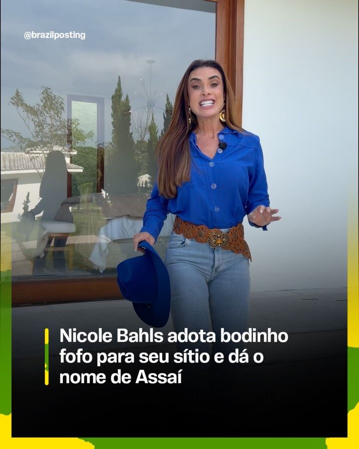 Mulher de cabelos castanhos e camisa azul em frente a janela de vidro, com vista para propriedade rural e jardinagem ao fundo.