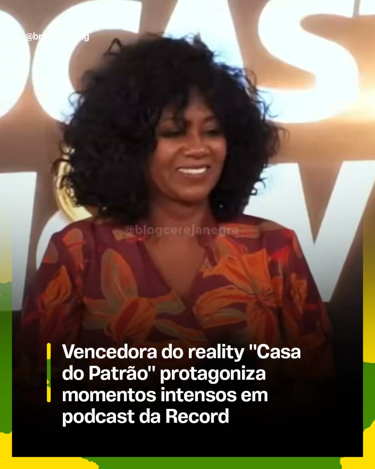 Mulher negra com cabelo liso preto em estúdio iluminado com letras brancas ao fundo, usando blusa de padrão laranja e vermelho, sorrindo para câmera