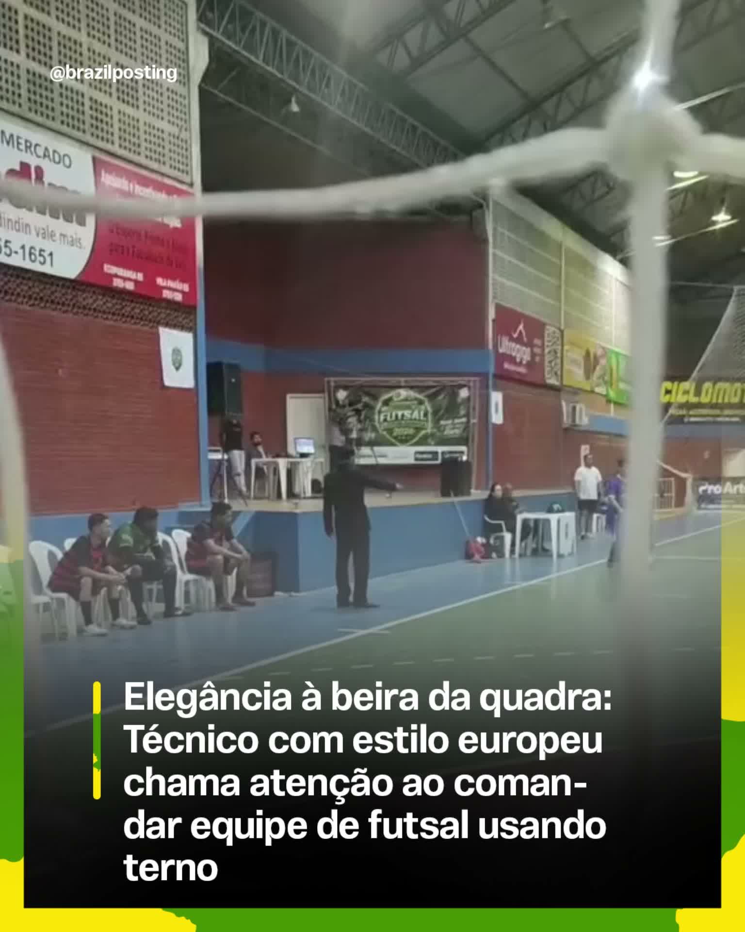 Técnico em terno e gravata em pé na lateral da quadra de futsal, observando jogo com jogadores em uniforme esportivo ao fundo.