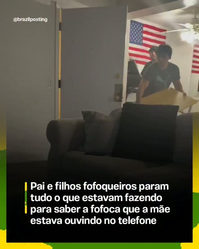 Homem de camiseta preta em sala de estar com sofá cinzento, bandeiras dos EUA ao fundo, observando com curiosidade uma conversa de telefone na parede ao lado