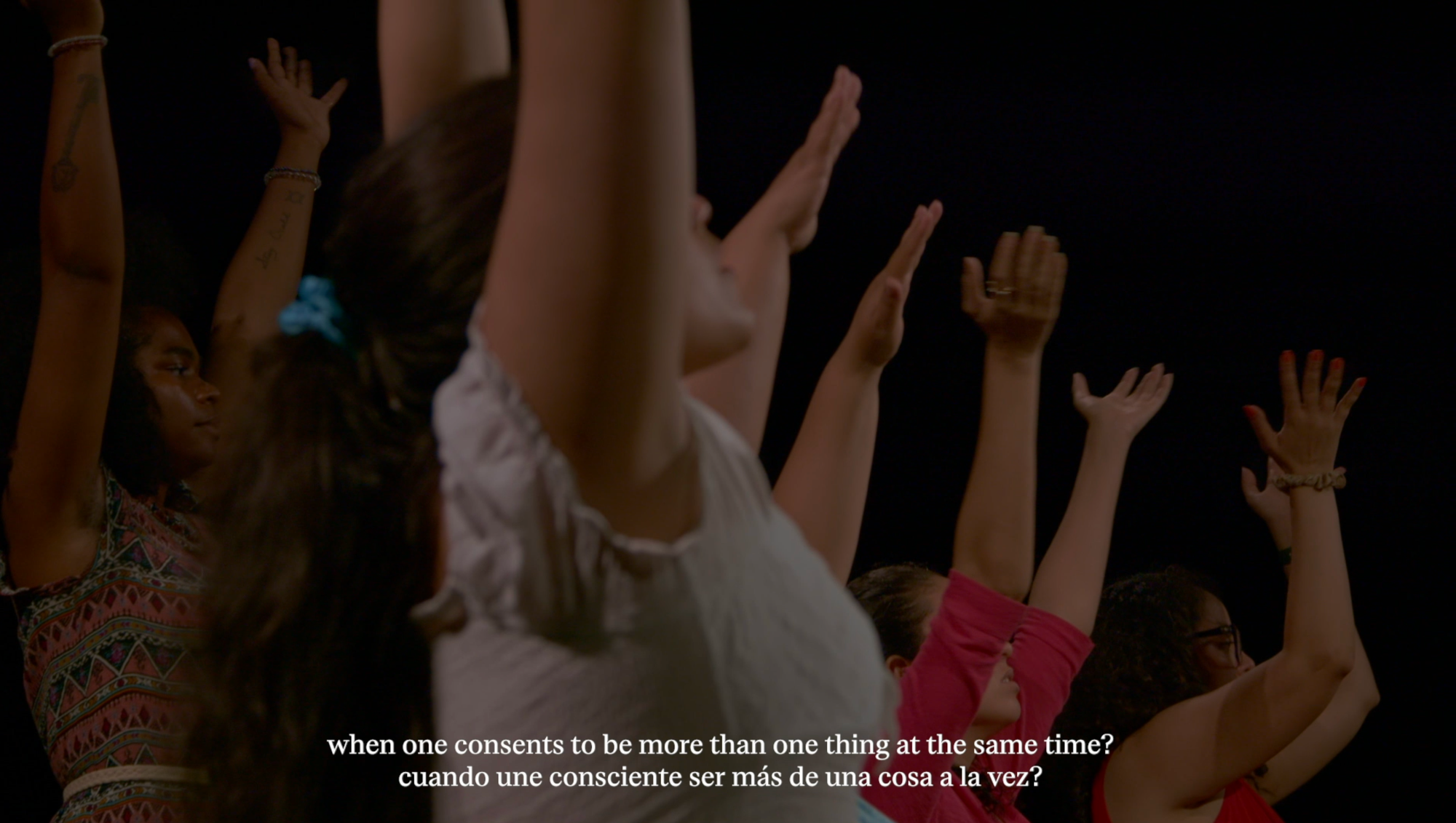 A film still where women stretch their arms above their heads, with a subtitle that reads: "when one consents to be more than one thing at the same time?" in English and Spanish. 