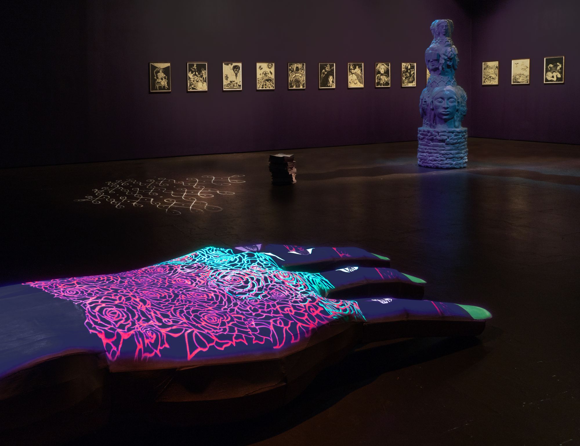 A massive sculptural hand in the foreground glows neon-like, while behind it is a room of work all tinged blue.