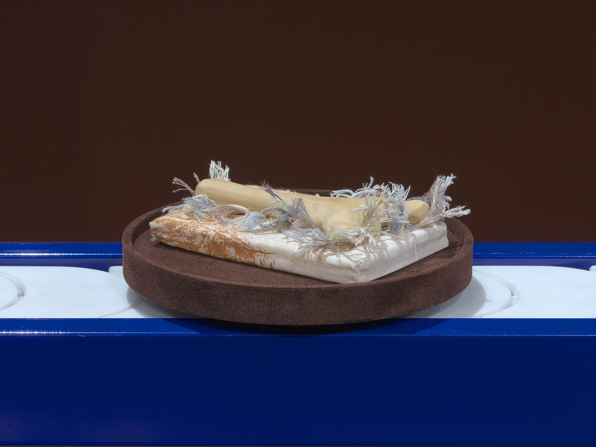 An embroidered pillow featuring a bonelike object, resting on a brown plate which moves along a blue conveyor belt.