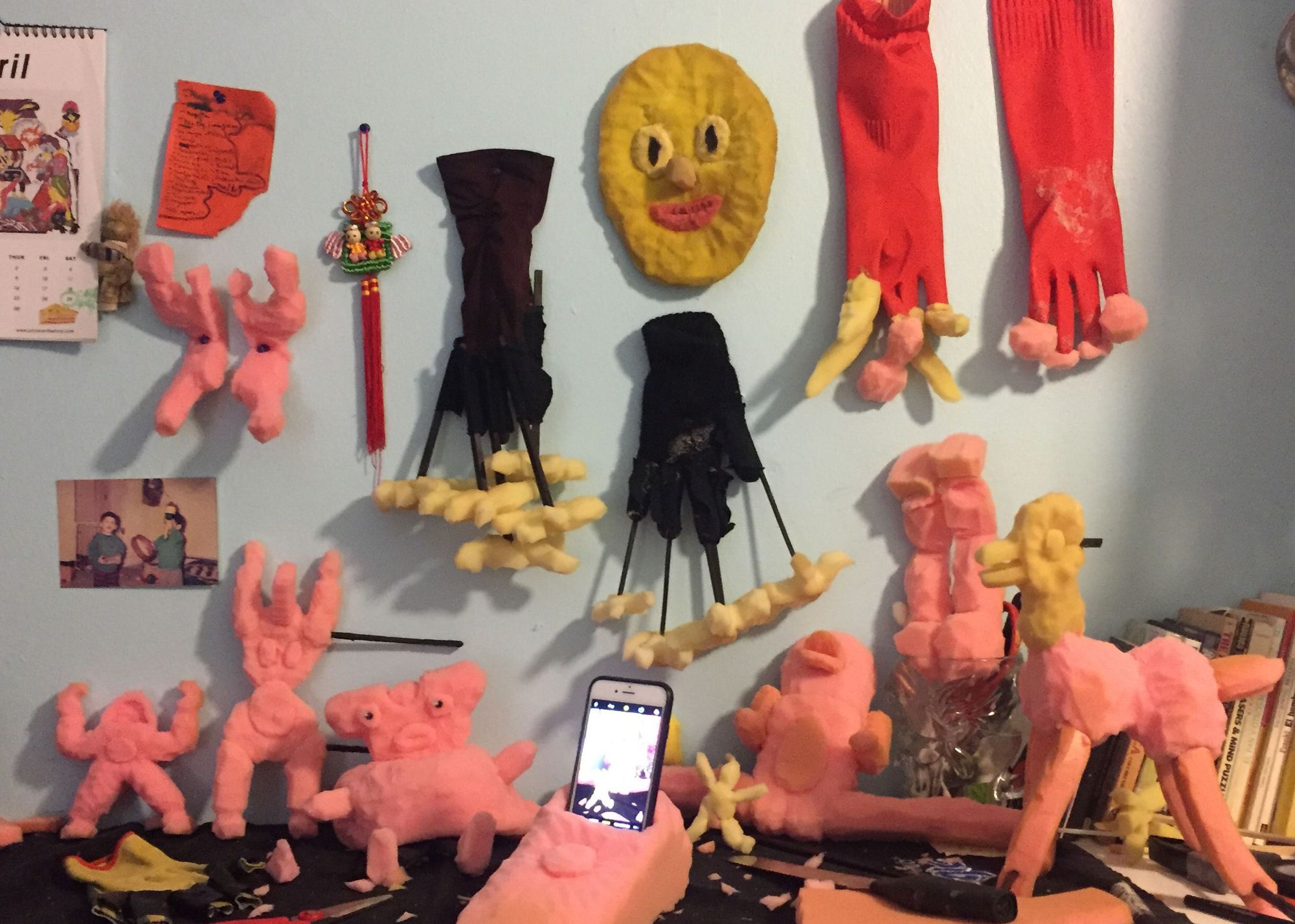 A desk covered in pink foam puppets and a cellphone, with performance gloves, puppets, and pictures hanging on the wall above it