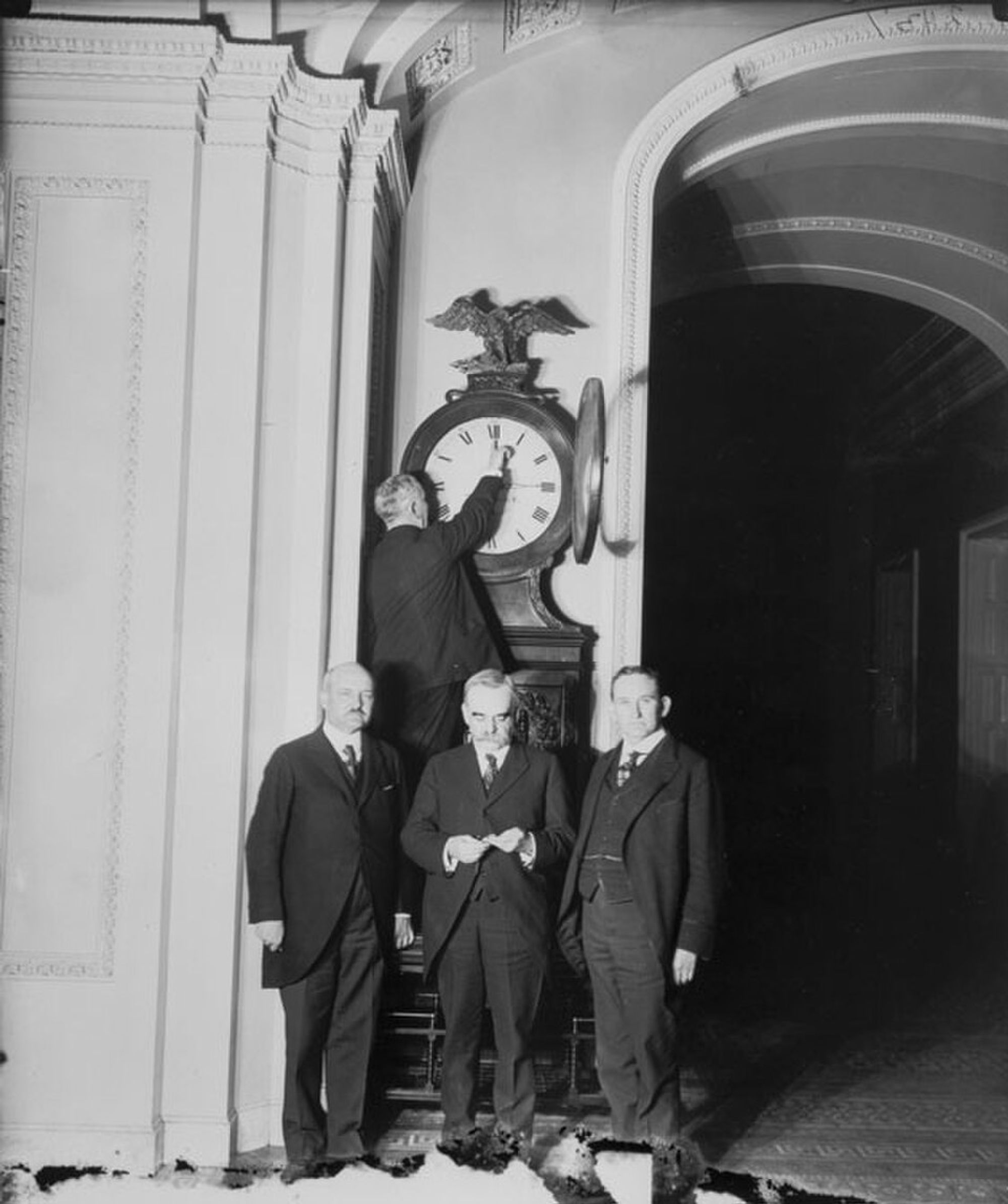 Black and white photograph. One man adjusts a large grandfather clock, and three other men stand and look at the camera. They are all white and wear suits.