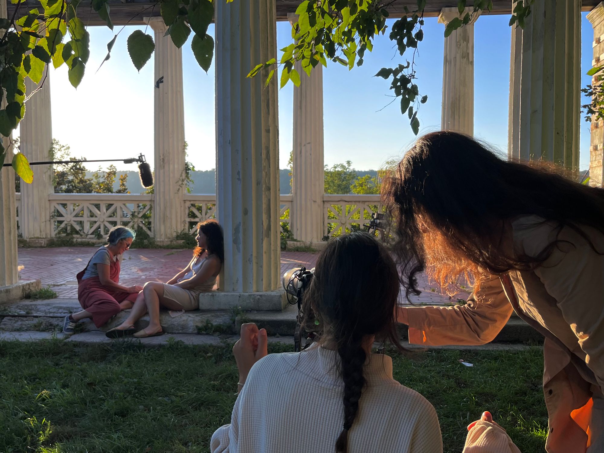 Two figures adjust a camera in the foreground, which focuses on two other figures sitting at some sort of veranda. A microphone floats above their heads.