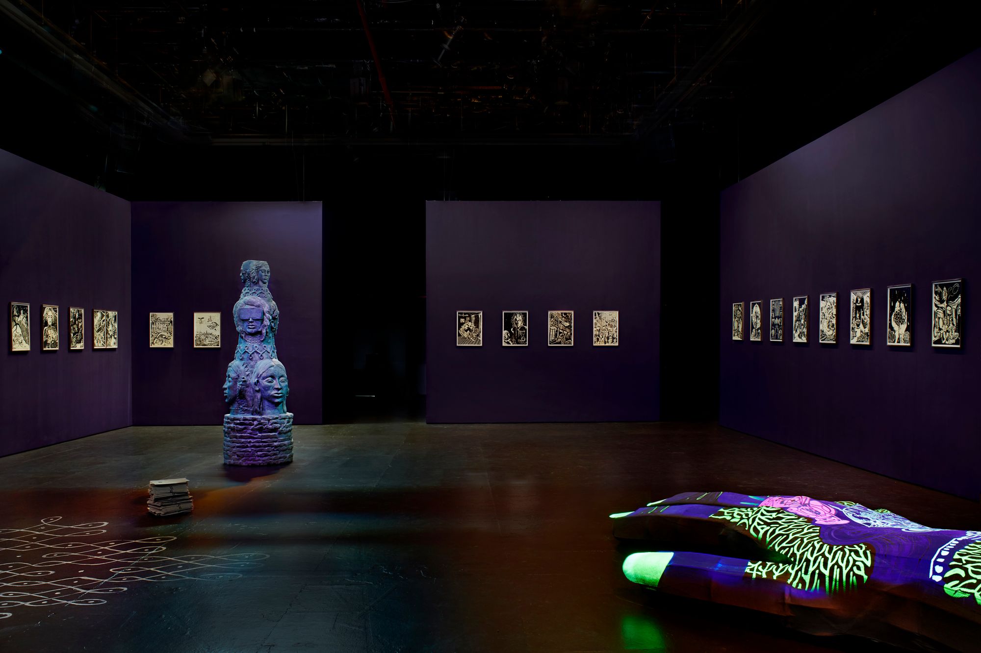 A wide-angle shot of Ganesh's exhibition "Her garden, a mirror," which shows a straight row of glowing prints on all surrounding walls, plus sculptures in the middle, also glowing blue.