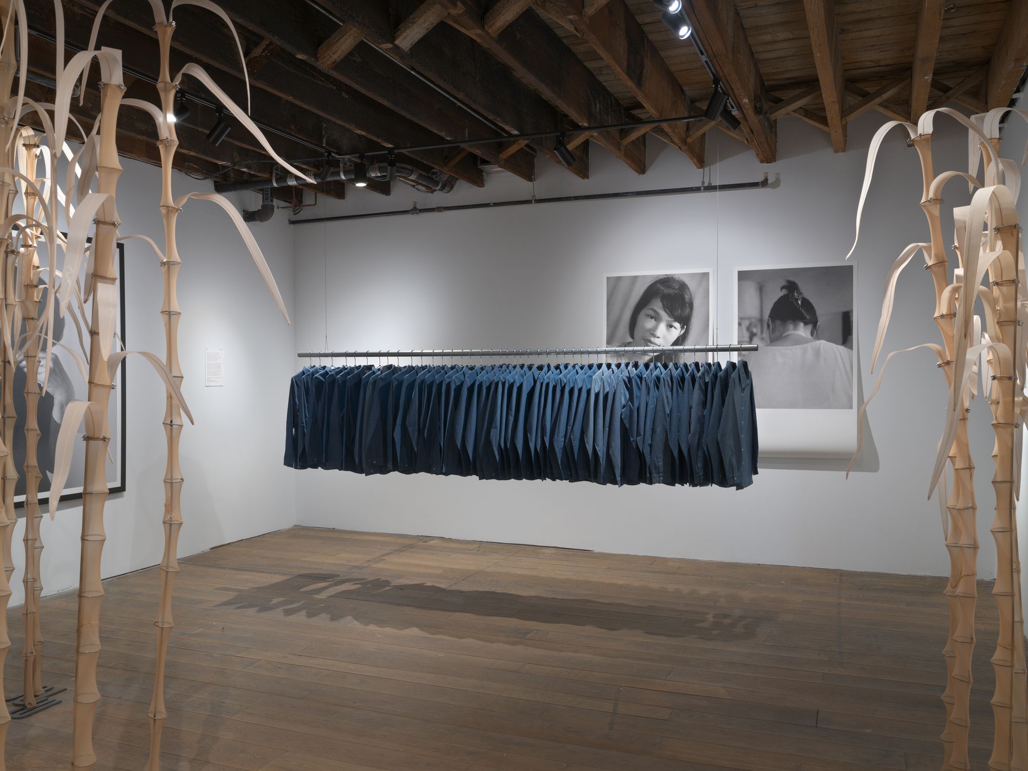 A gallery space featuring a collection of denim shirts hanging from a free-floating rack, with two black-and-white images on the wall behind them and artificial sugar cane stalks rising from the floor in the foreground.
