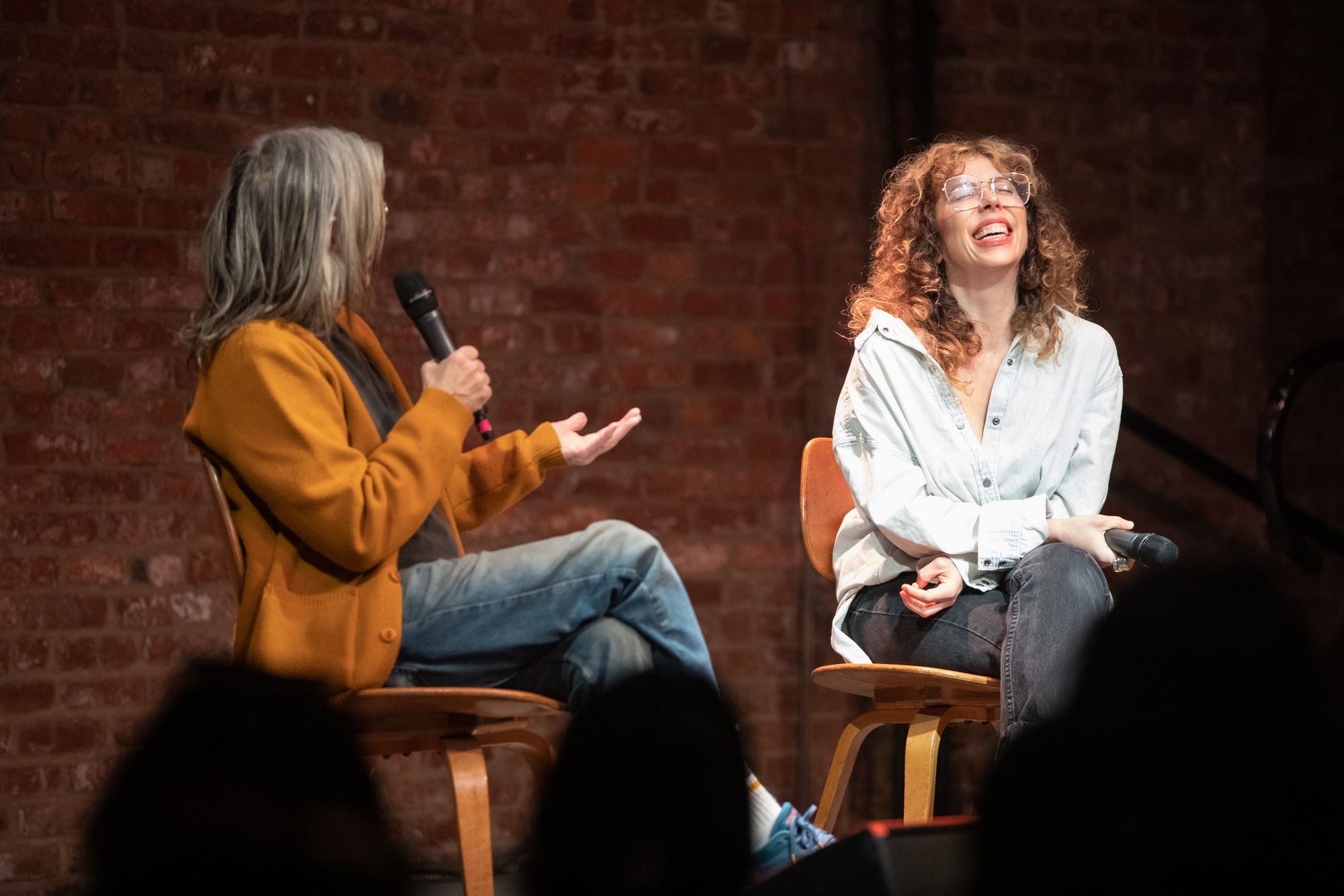 Eileen Myles speaks into a microphone, facing towards Ariana Reines who's tilting her head back with laughter.