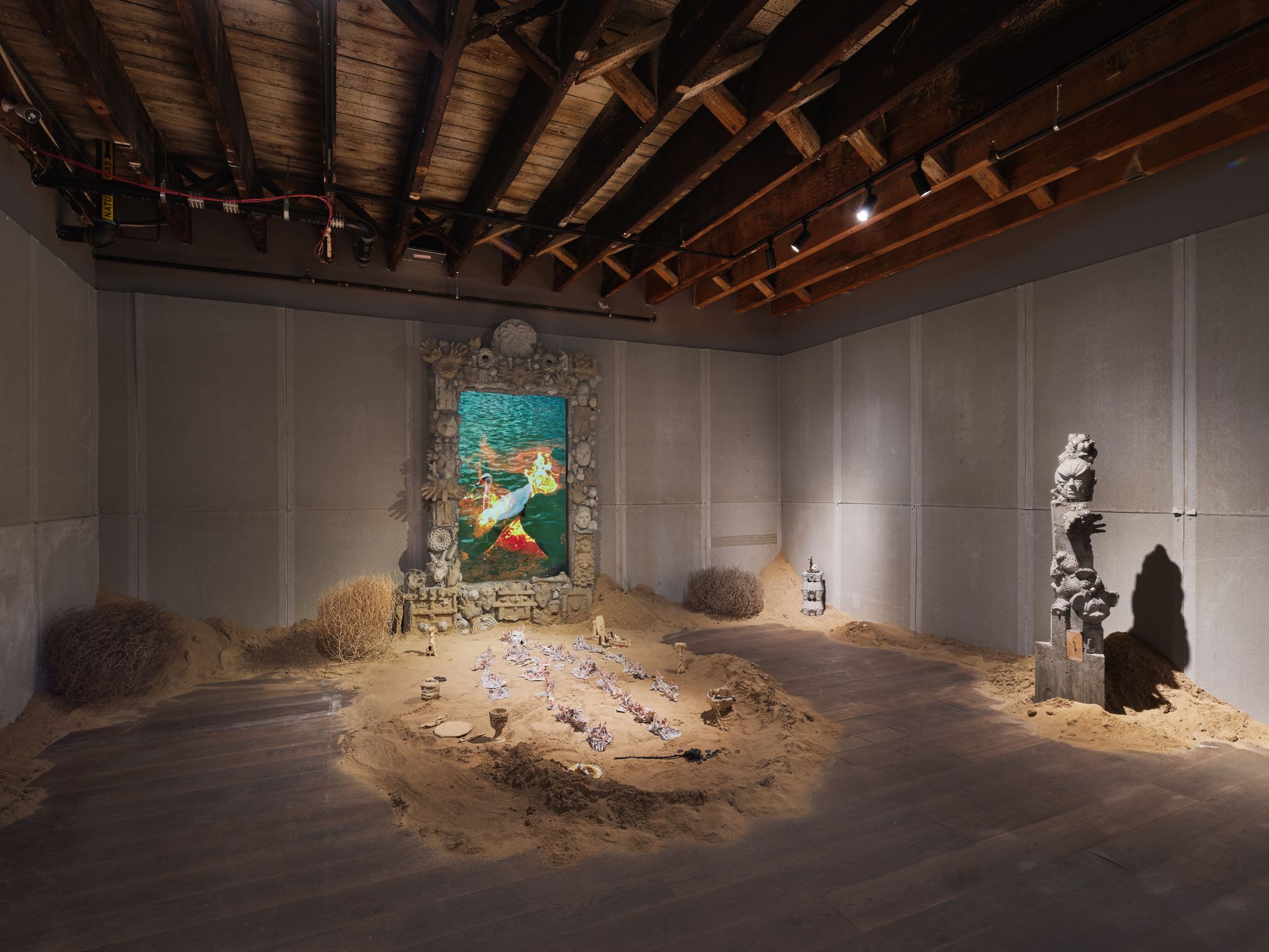 A low-lit gallery space wherein a screen decorated in shells is propped atop a bed of sand, covered in ceramic trinkets, and totems decorate the other walls.