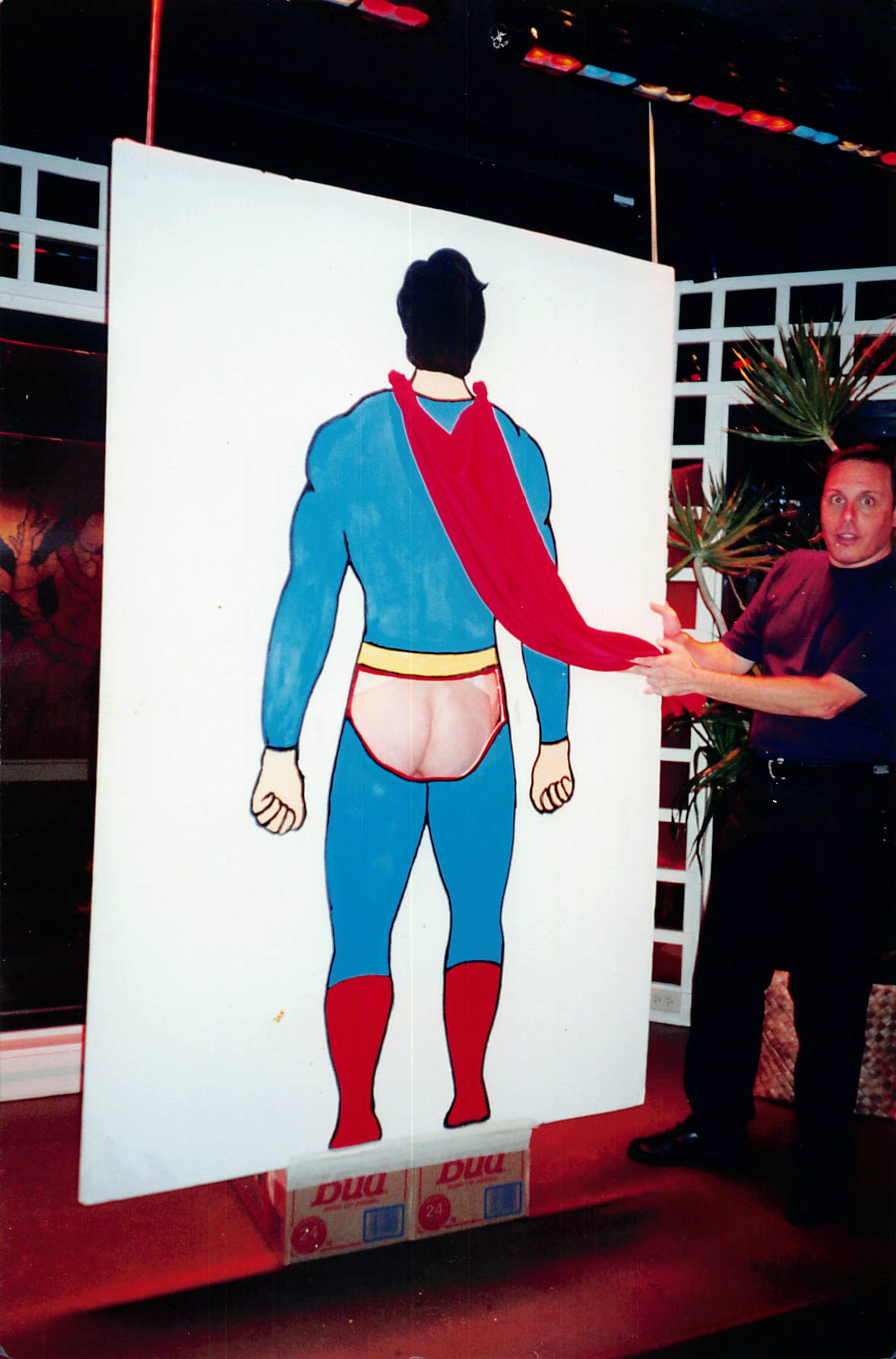 A man holds the cape that hangs off of a wooden cut-out of Superman's backside, exposing a bare ass exposed where the character's red briefs would be.