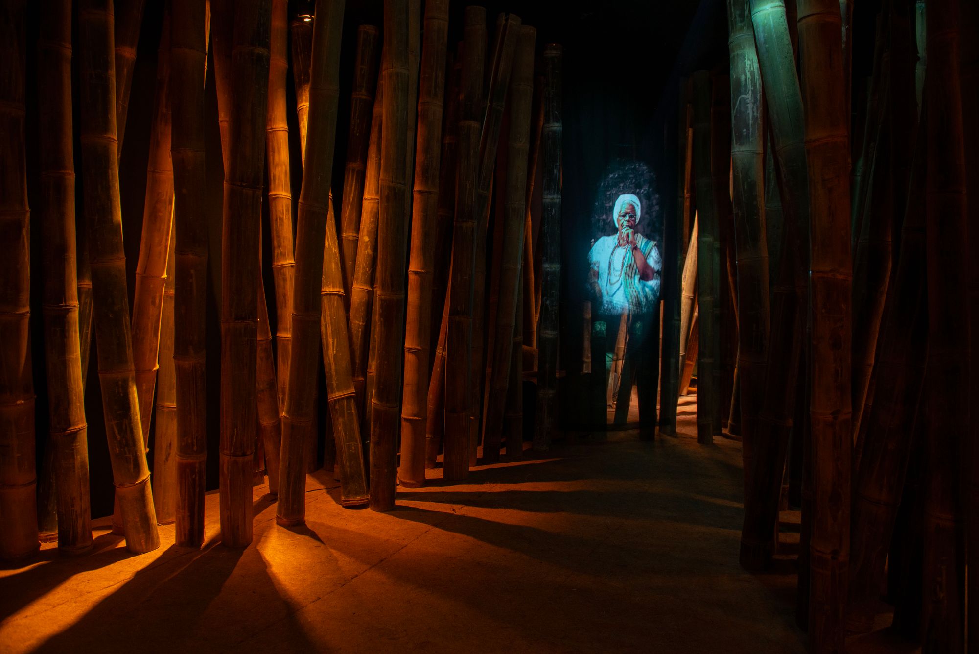 Soft orange light spills out from behind bamboo shoots in a dark room; a screen lights the right side with the image of a black woman bathed in sky blue light.