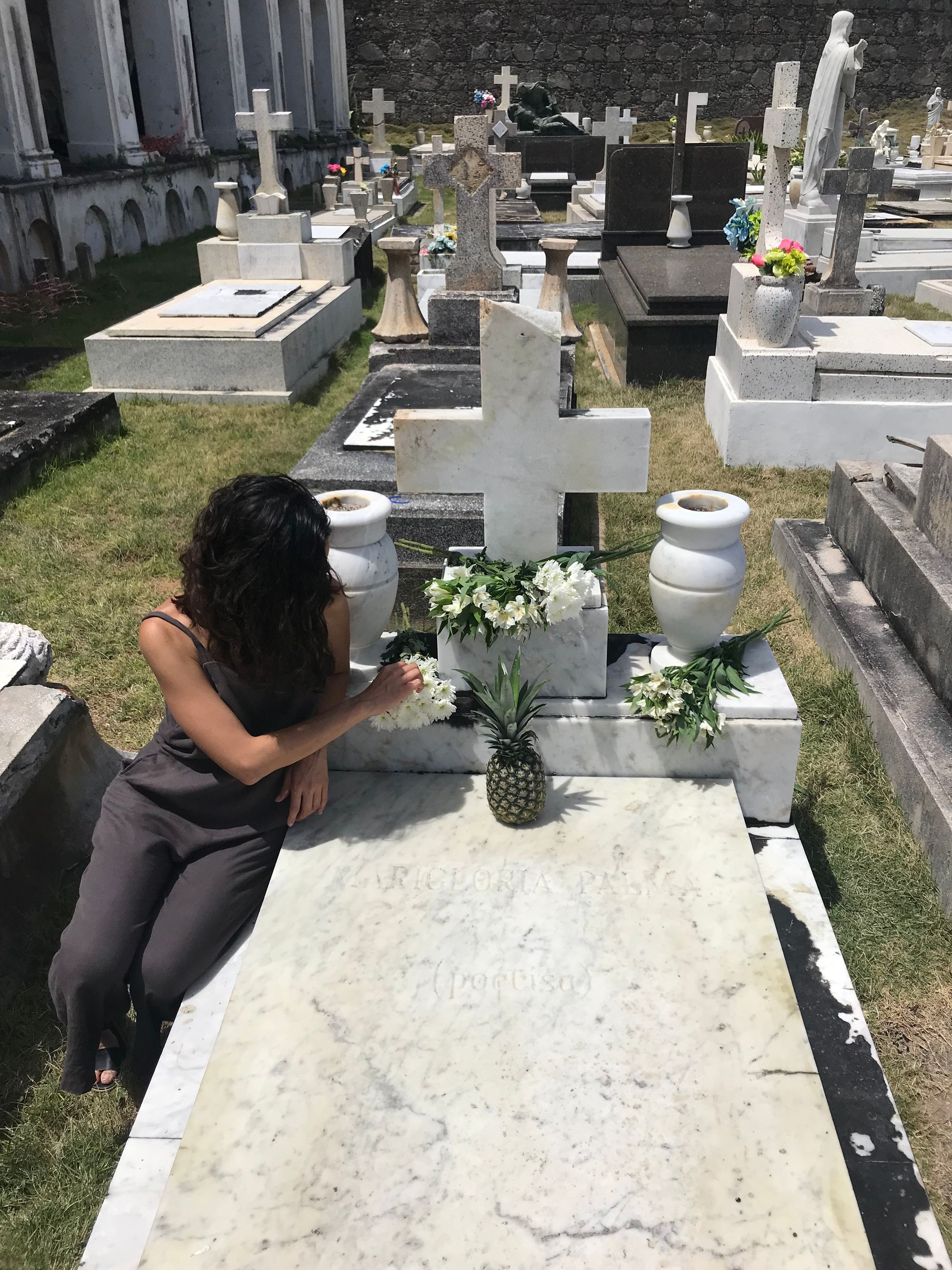 A woman sits by a white marble tombstone, her face covered by her own dark hair as she turns to her left and adjusts the flowers she's laid out.