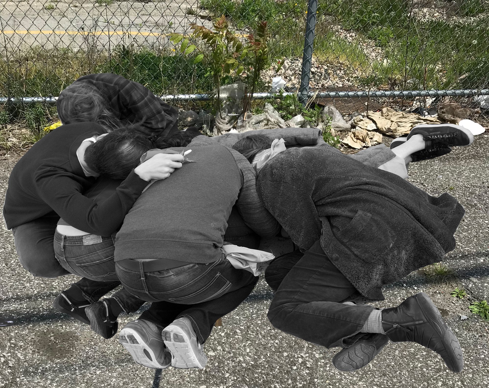 a group of bodies, some crouching and slouched over one another on asphalt with a chain link fence in the background