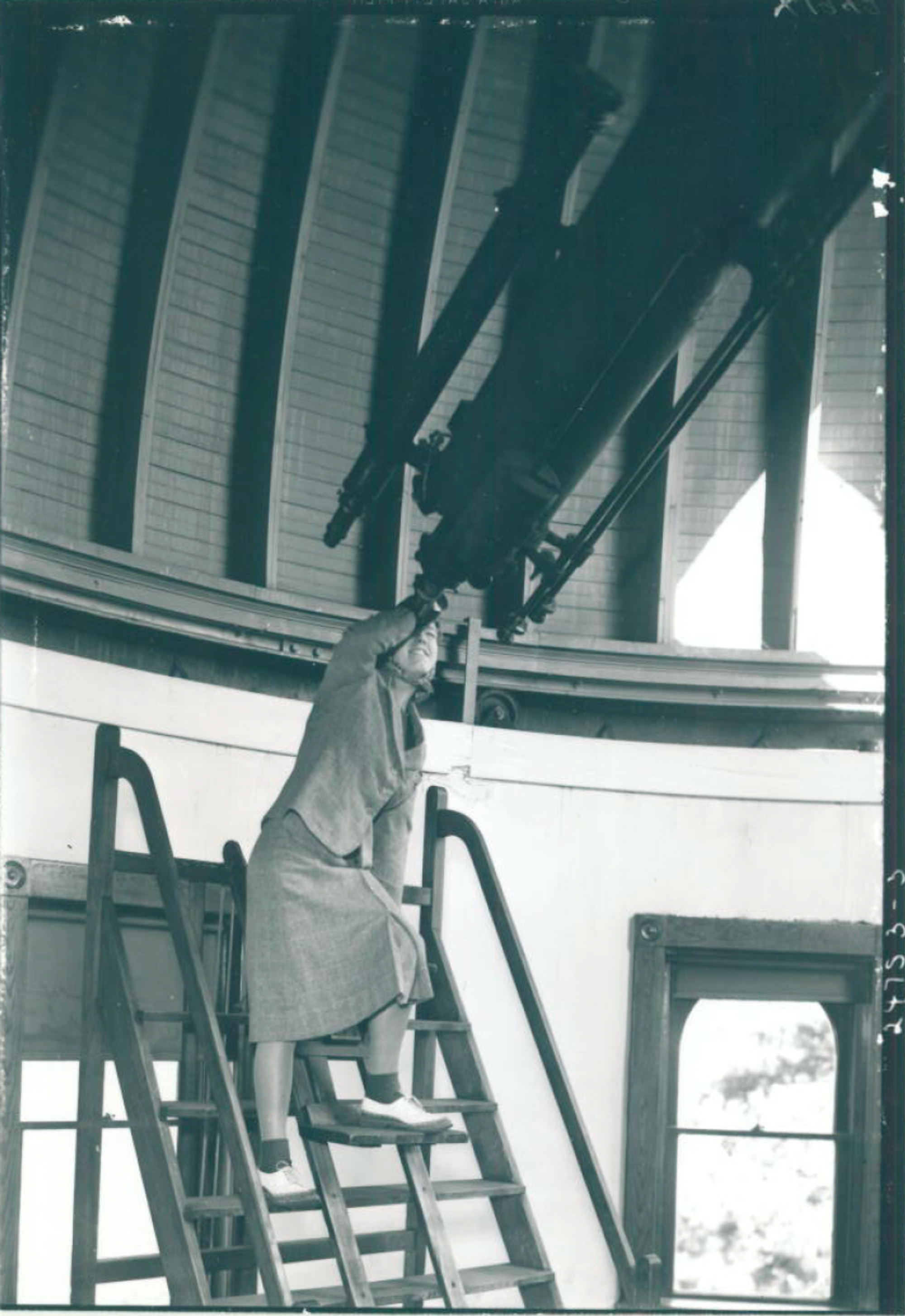 A black and white picture of a woman standing awkwardly on metal stairs as she peers into a massive telescope.