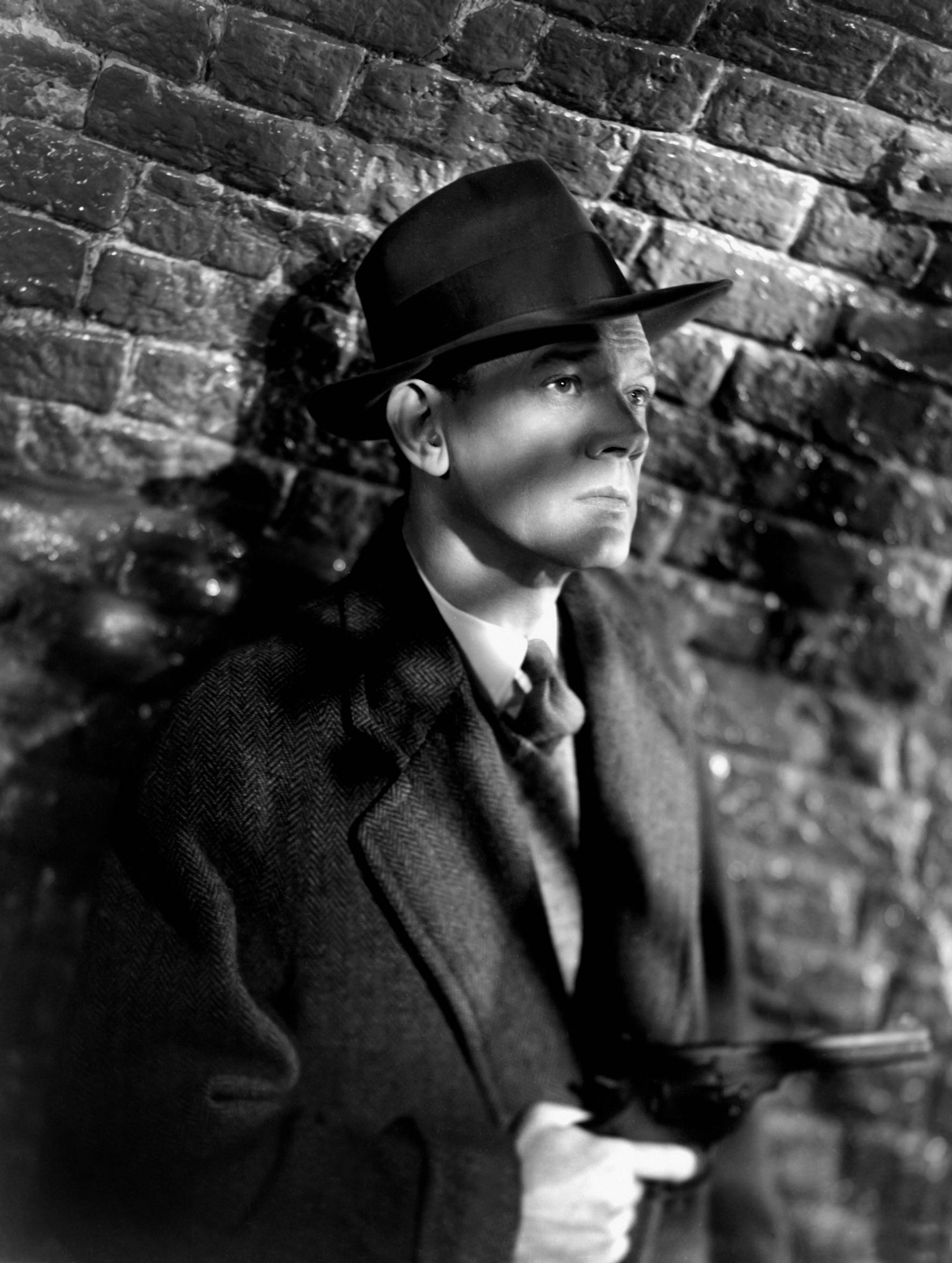 A black-and-white photograph of a man in a suit and a top hat standing against a brick wall.