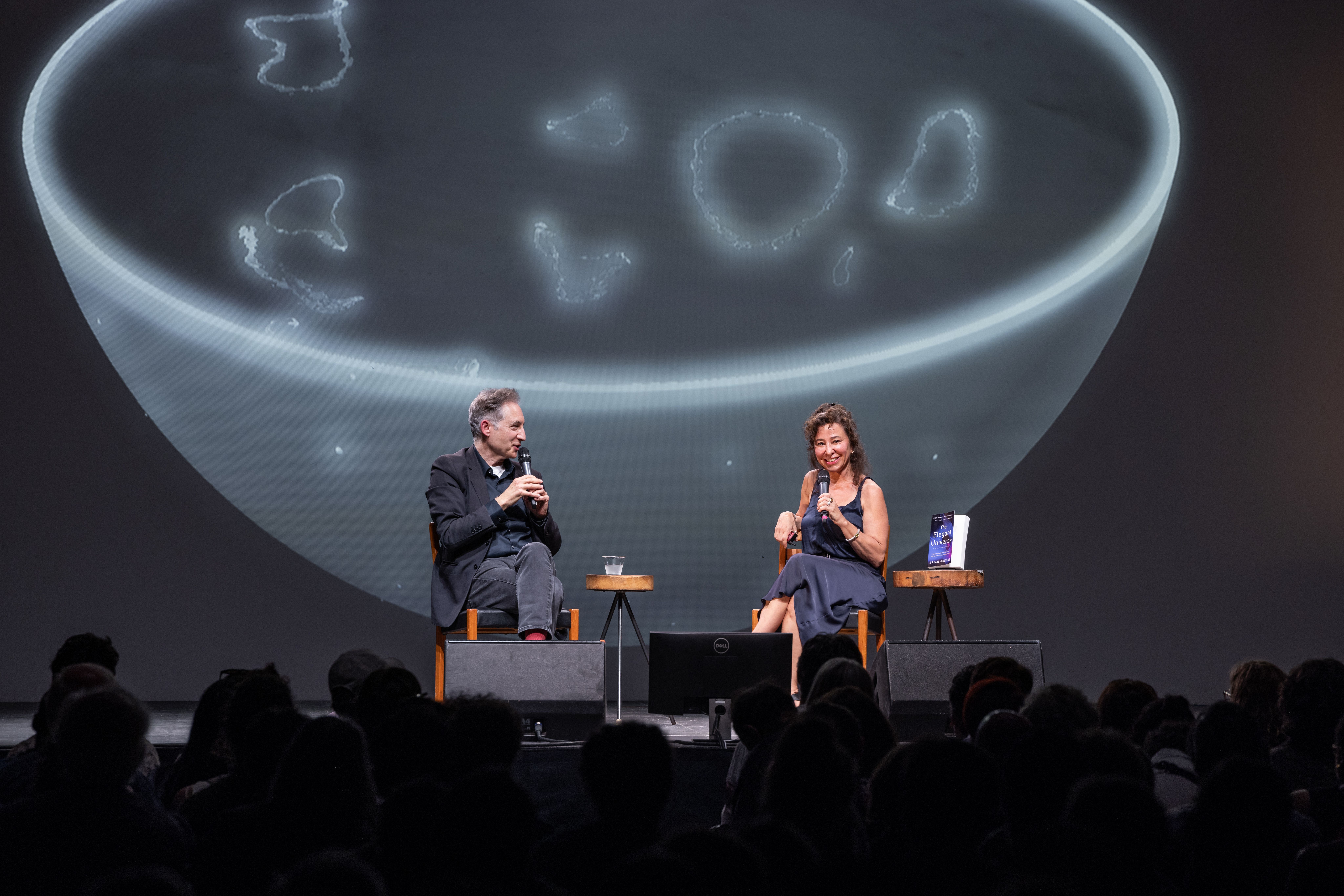 Brian Greene and Janna Levin chat on stage, smiling into their microphones, against a projected backdrop resembling a glowing bowl