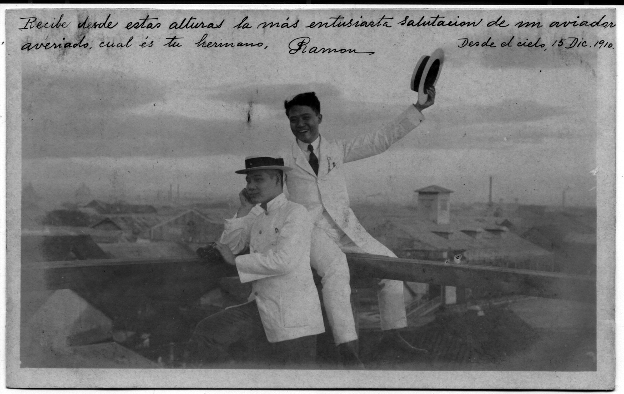 A black-and-white portraiture photograph where two men, Constancio and Ramón, are posed. One is wearing a hat and a collared double-breasted jacket and is sitting while looking to the left. Another man is sitting on a higher ledge and is wearing a buttoned up white jacket and tie. He holds his hat out raised above his head like a greeting. He is smiling with teeth. There is script above the photograph.