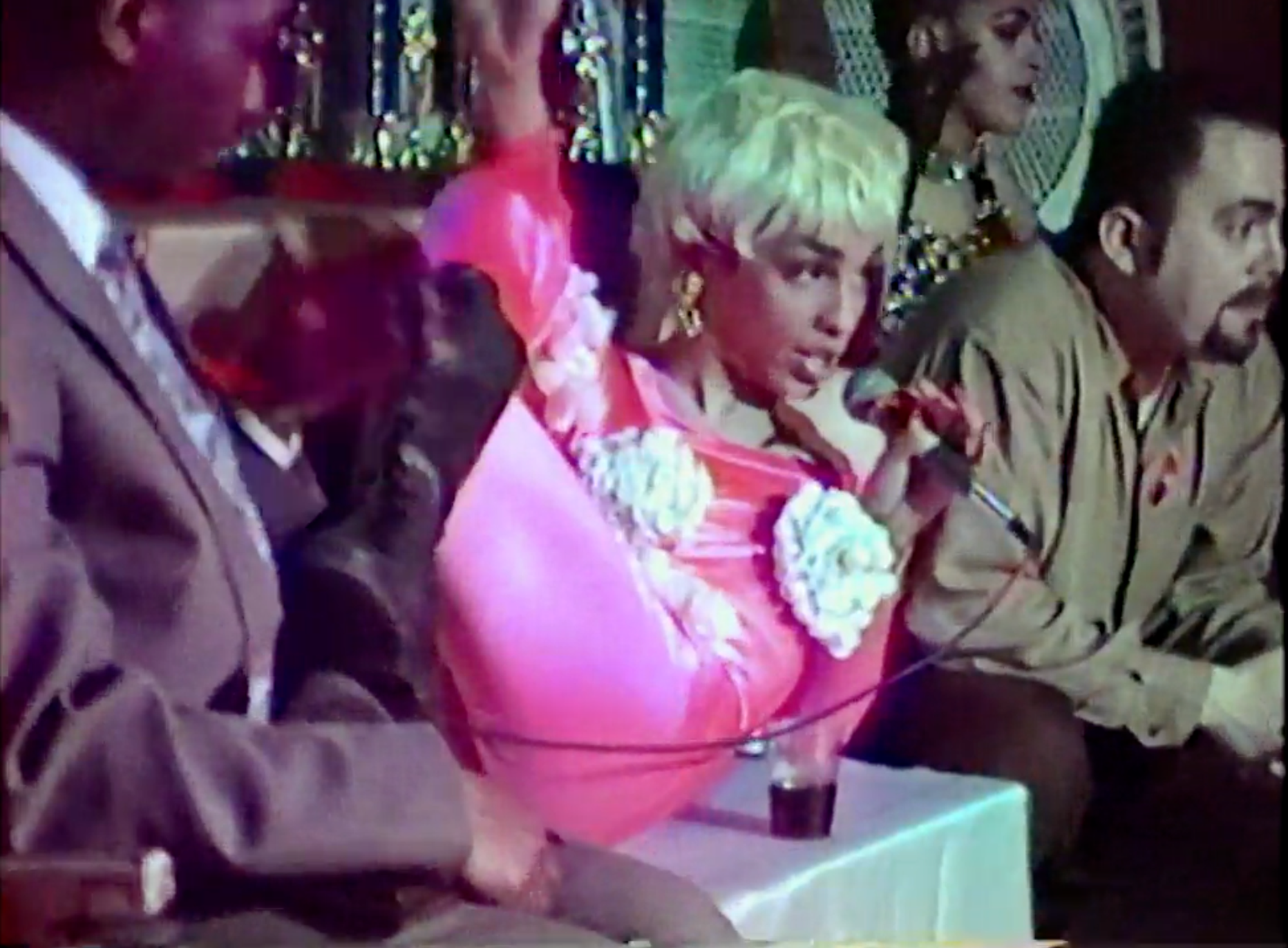A woman wearing a hot pink dress and white wig holding a mic laying across the stage