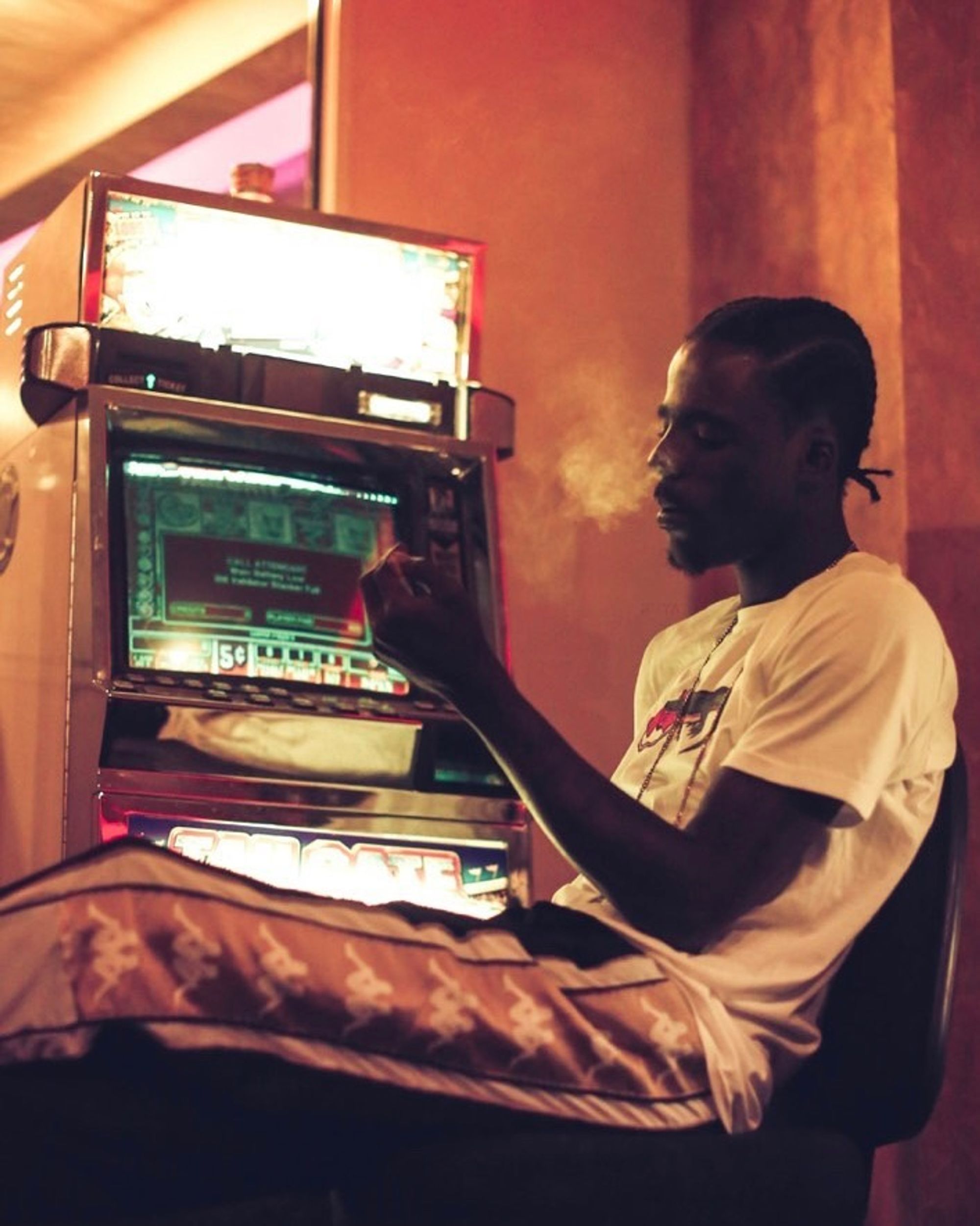 A color photograph of a man sitting in front of jukebox, smoking and looking into his own lap.