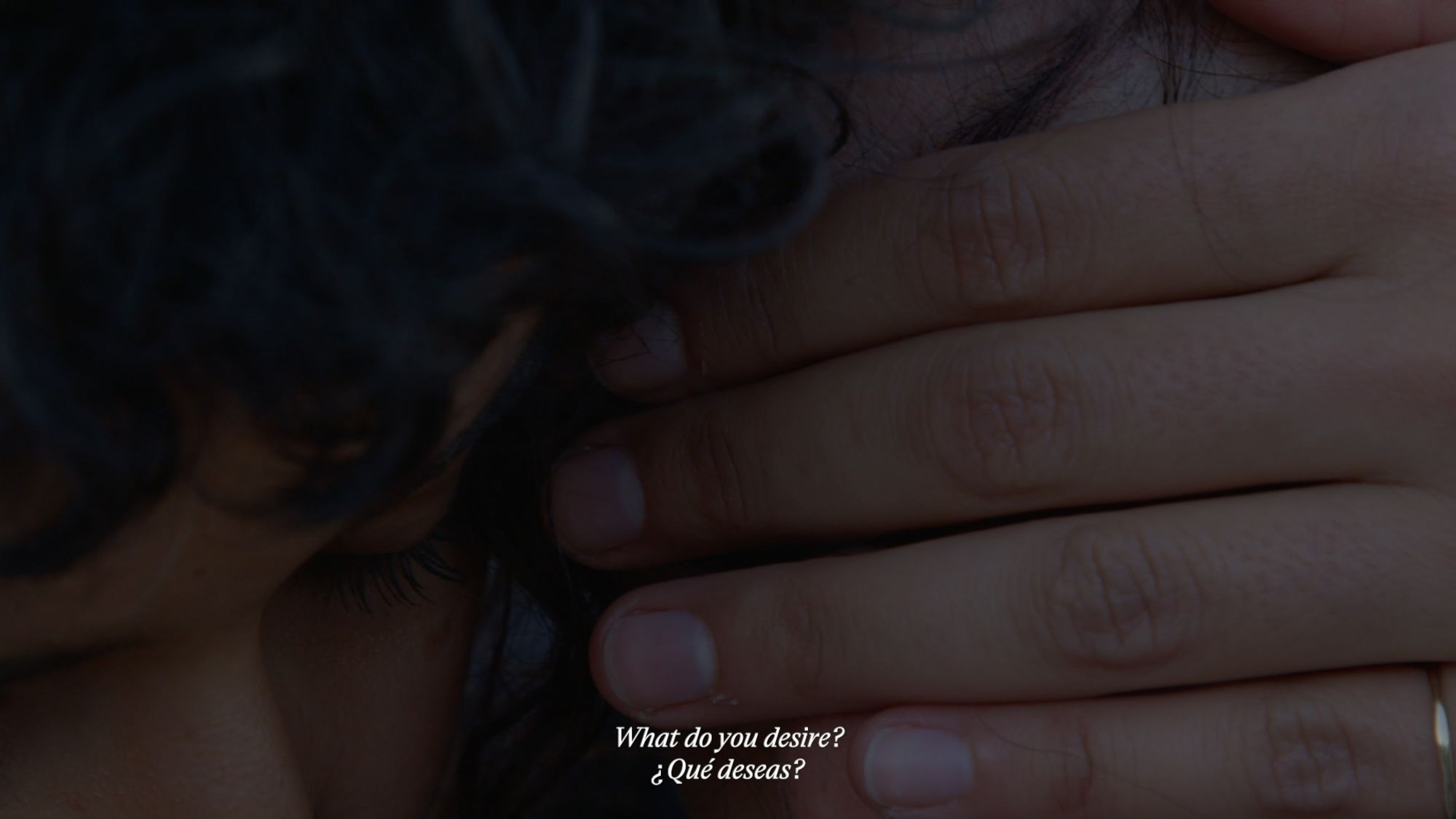 A film still of one person whispering to another, with their hand covering their face and the right half of the image. The subtitle reads: "What do you desire?" in both English and Spanish.