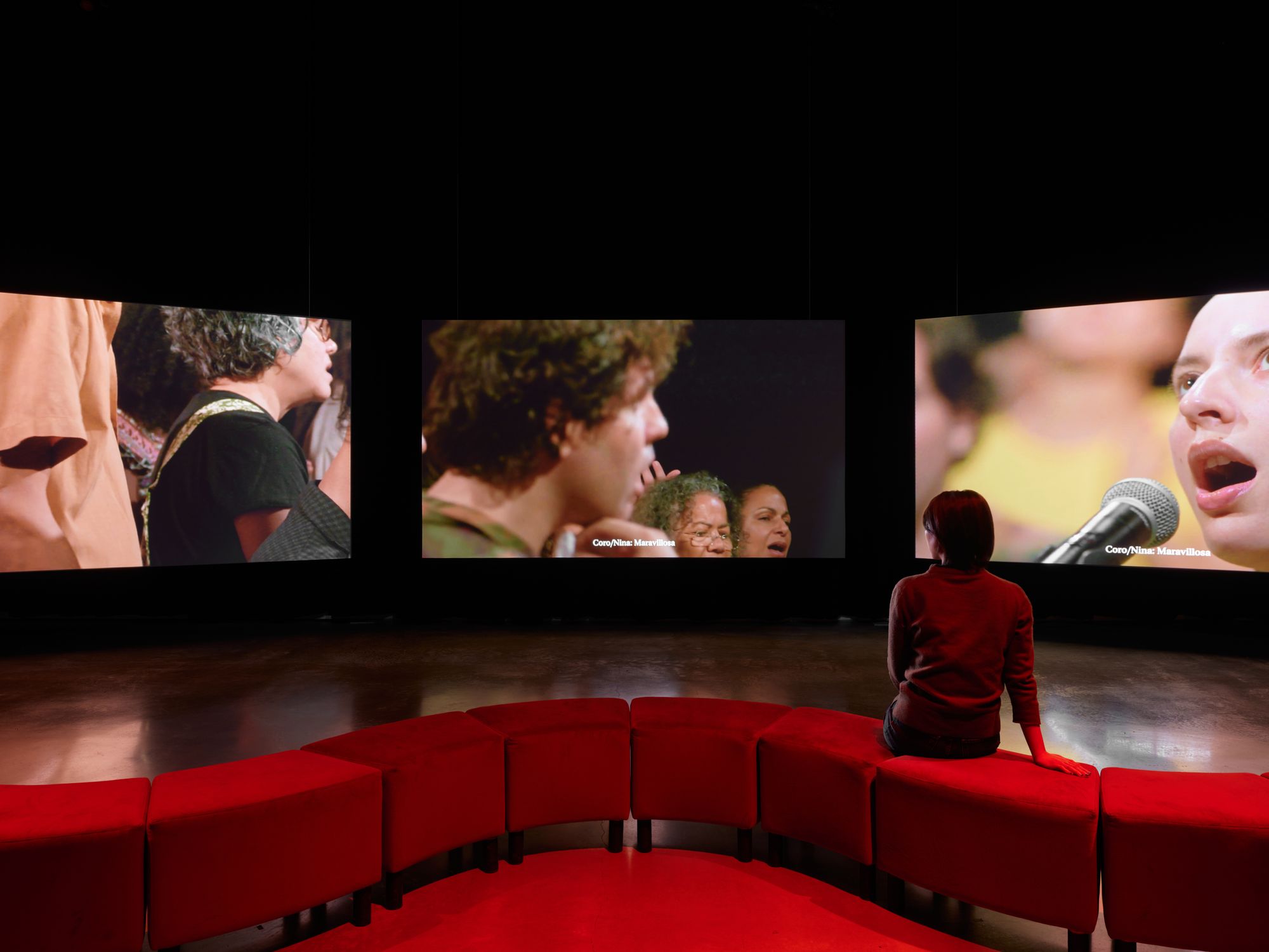 A figure sits on a red couch, staring at three screens in front of her which each showcase people at different angles.