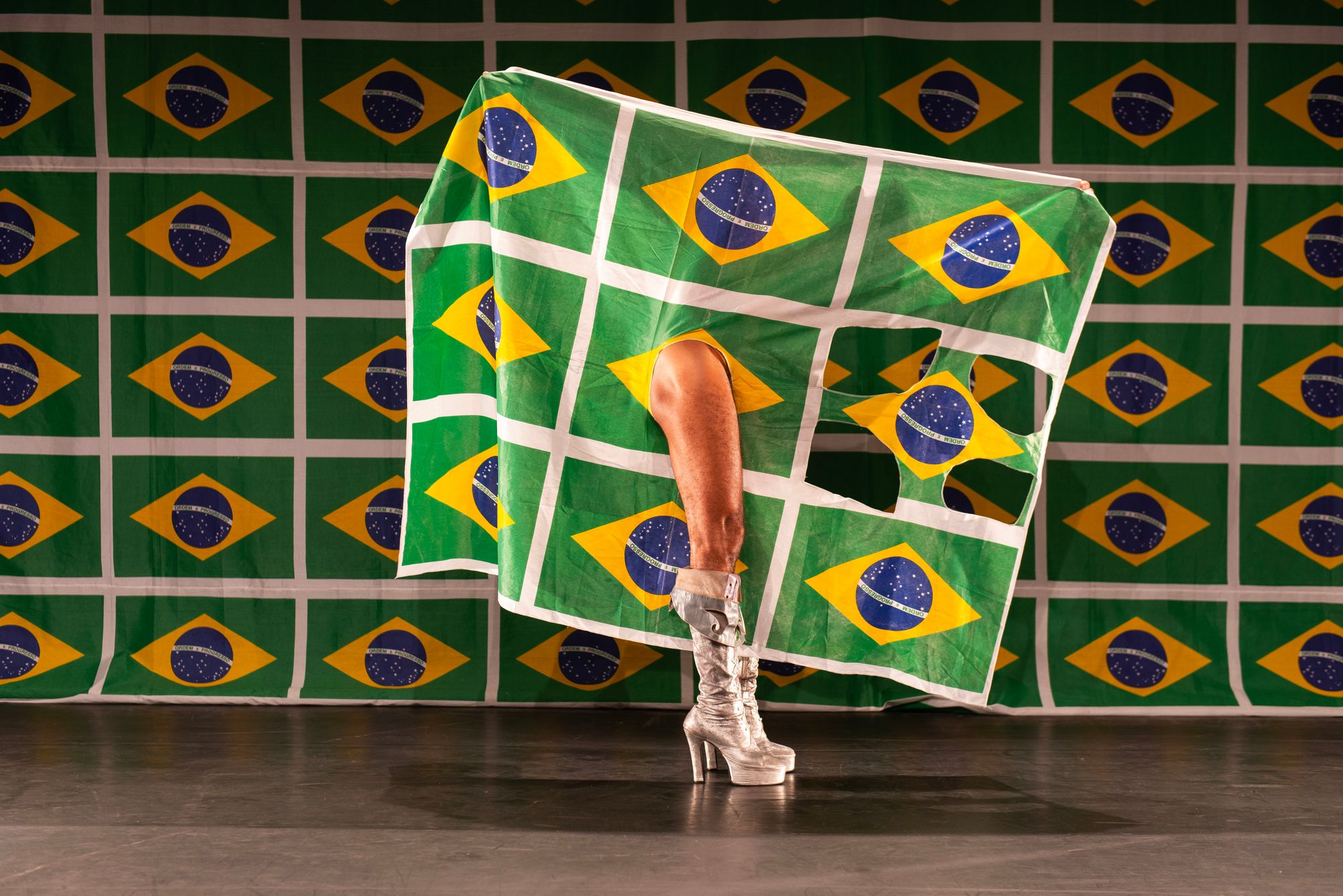 A muscular, stillettoed leg strikes through the Brazilian flag and stands in front of a backdrop where the flag is arranged in a gridlike formation.