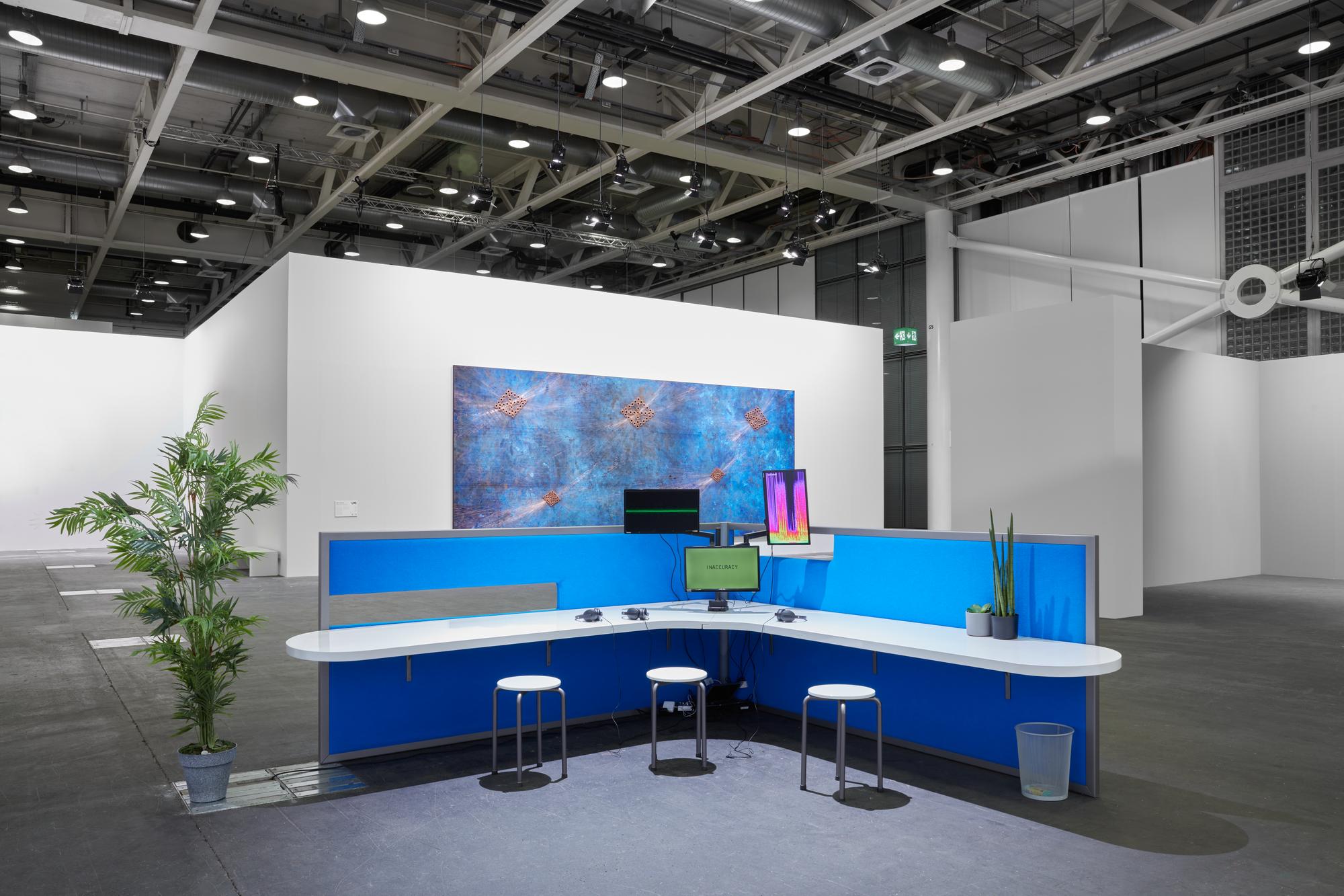 A view of Lawrence abu Hamdan's installation, of a blue office-space-like cordon that sections off a desk, monitors, and three stools. There is a spiky-leafed plant to the left side.