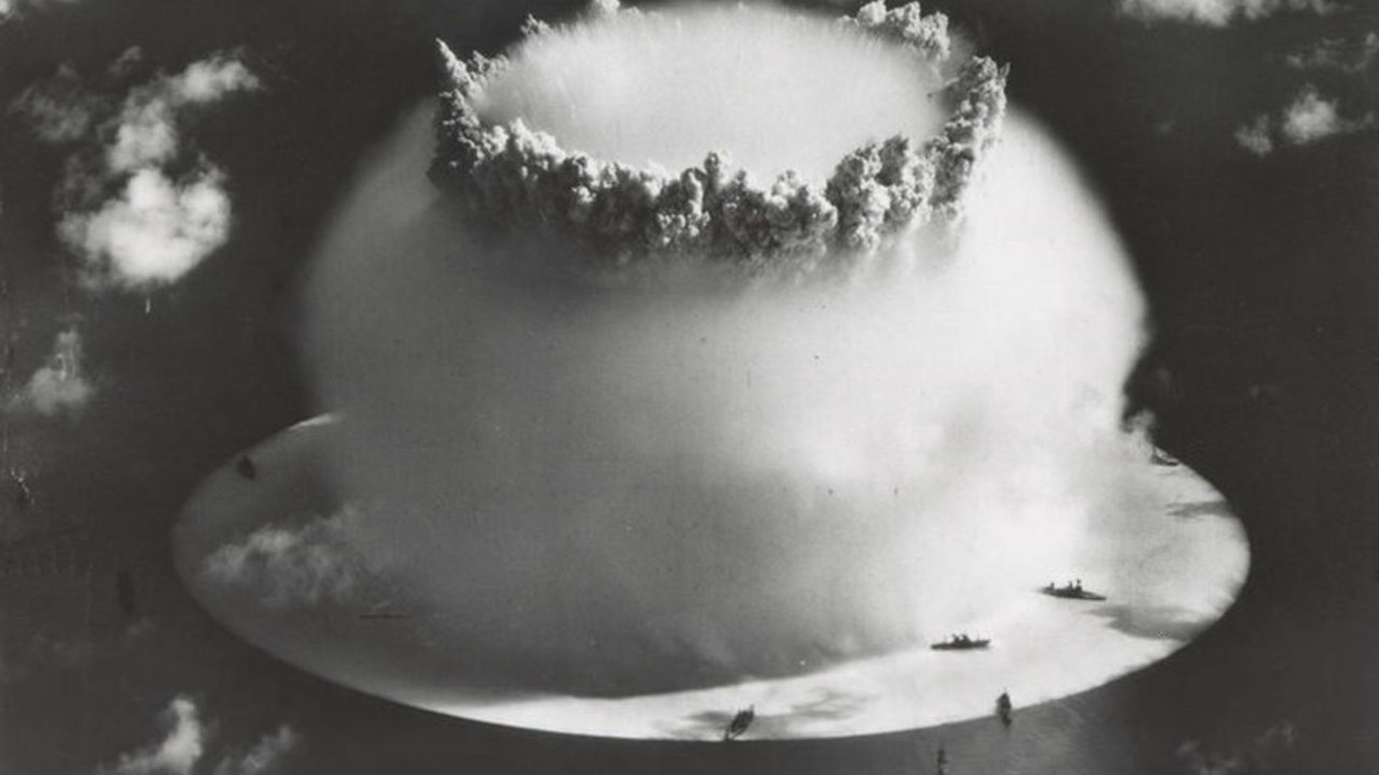 The Baker test, the second nuclear detonation of Operation Crossroads at Bikini Atoll, showing the white surface crack under the ships, and the top of the hollow spray column protruding through the hemispherical Wilson cloud.